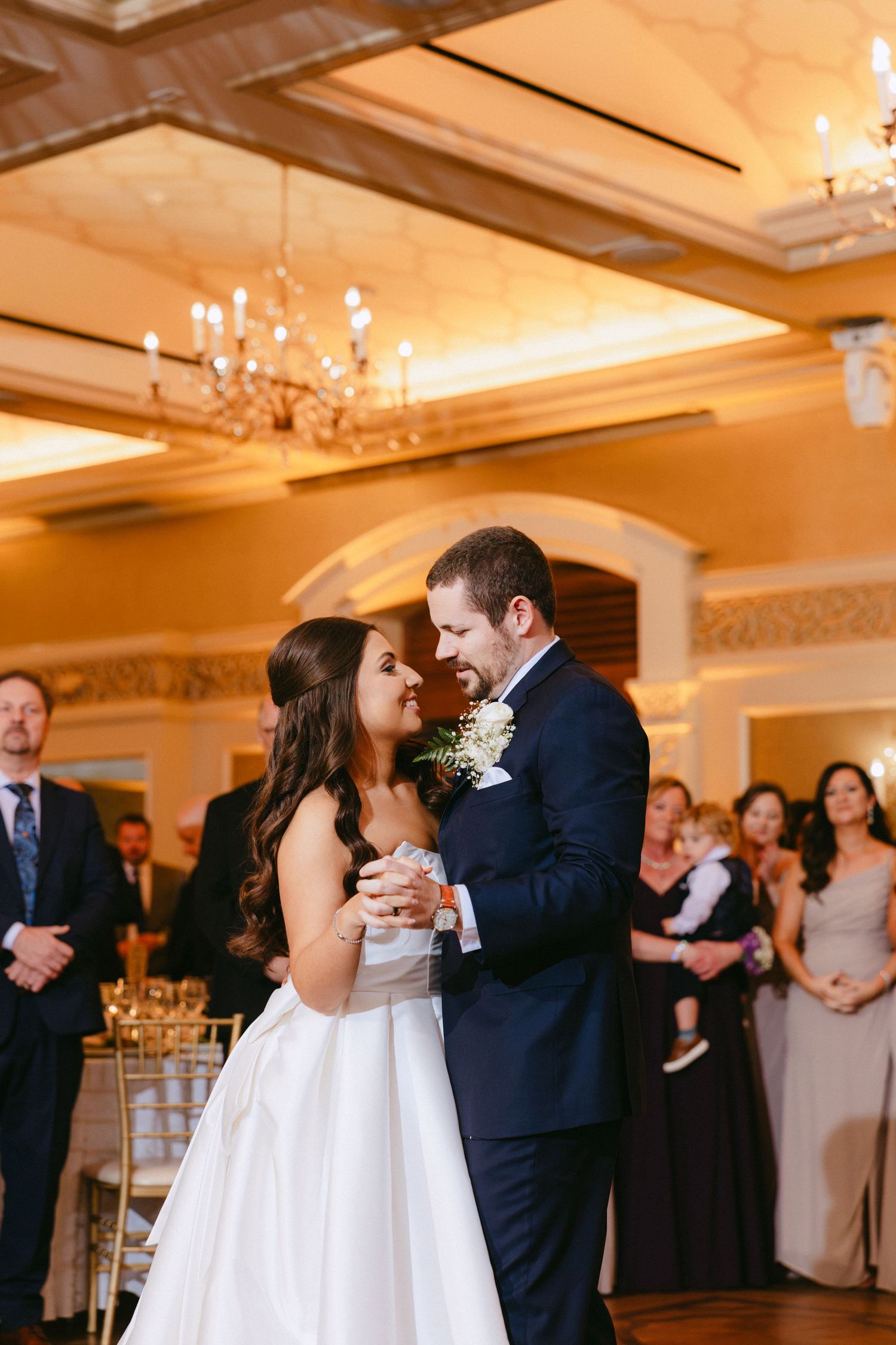 Bride and groom share first dance in elegant chandelier-lit ballroom surrounded by wedding guests.