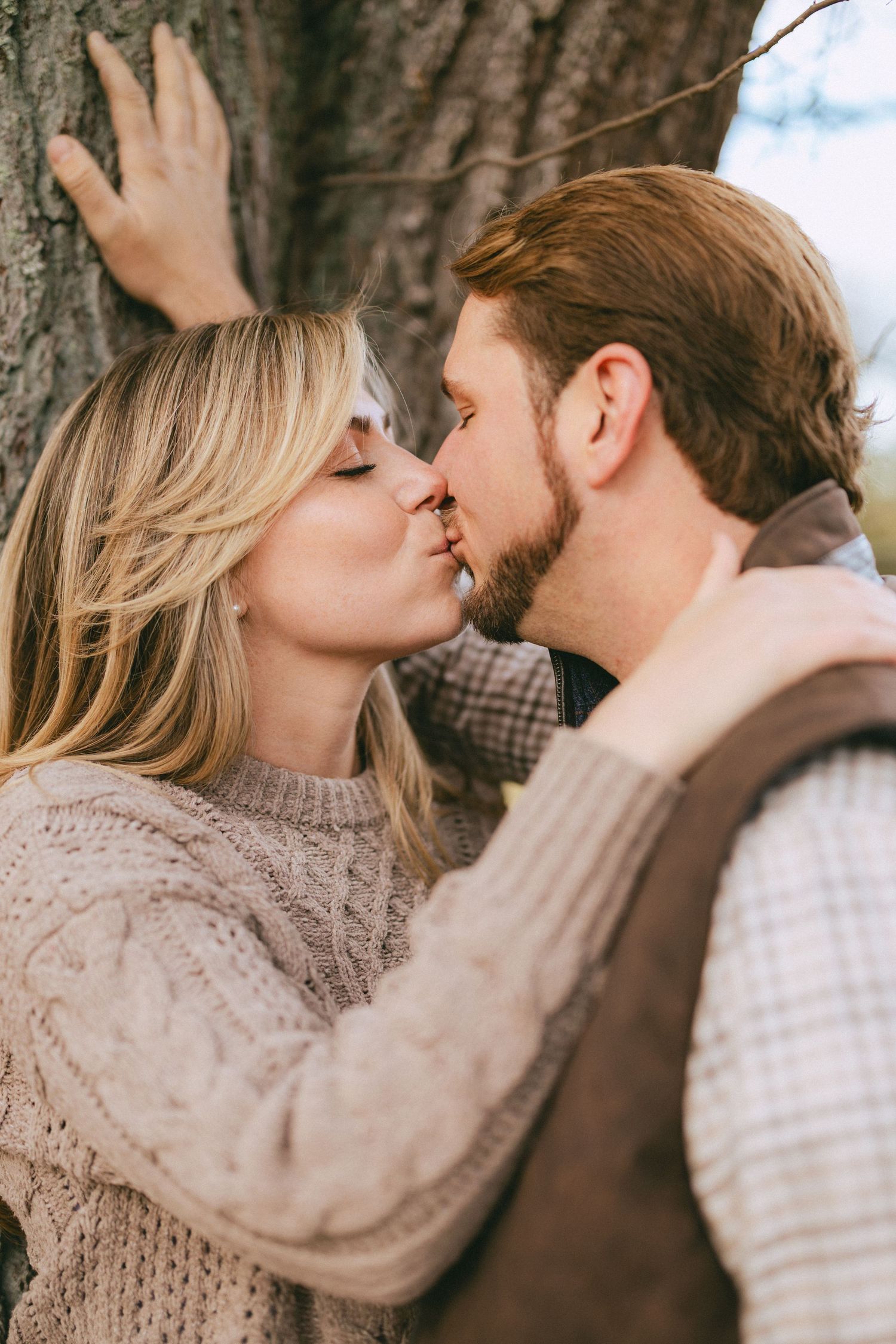 Couple shares a romantic kiss beside a large tree trunk in an outdoor fall setting.