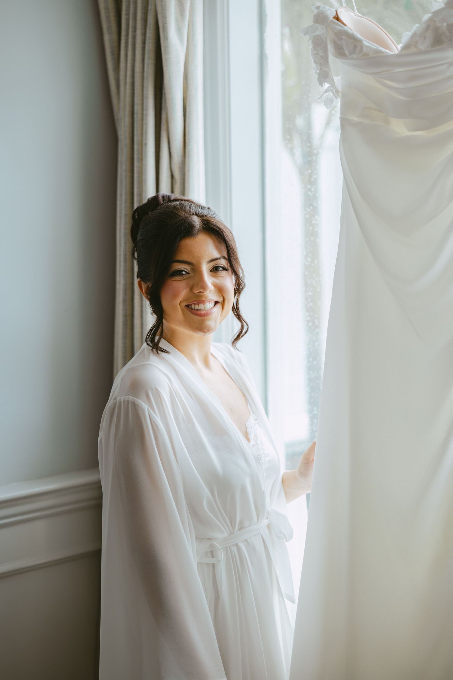 Smiling bride in white robe standing by window next to her hanging wedding dress.