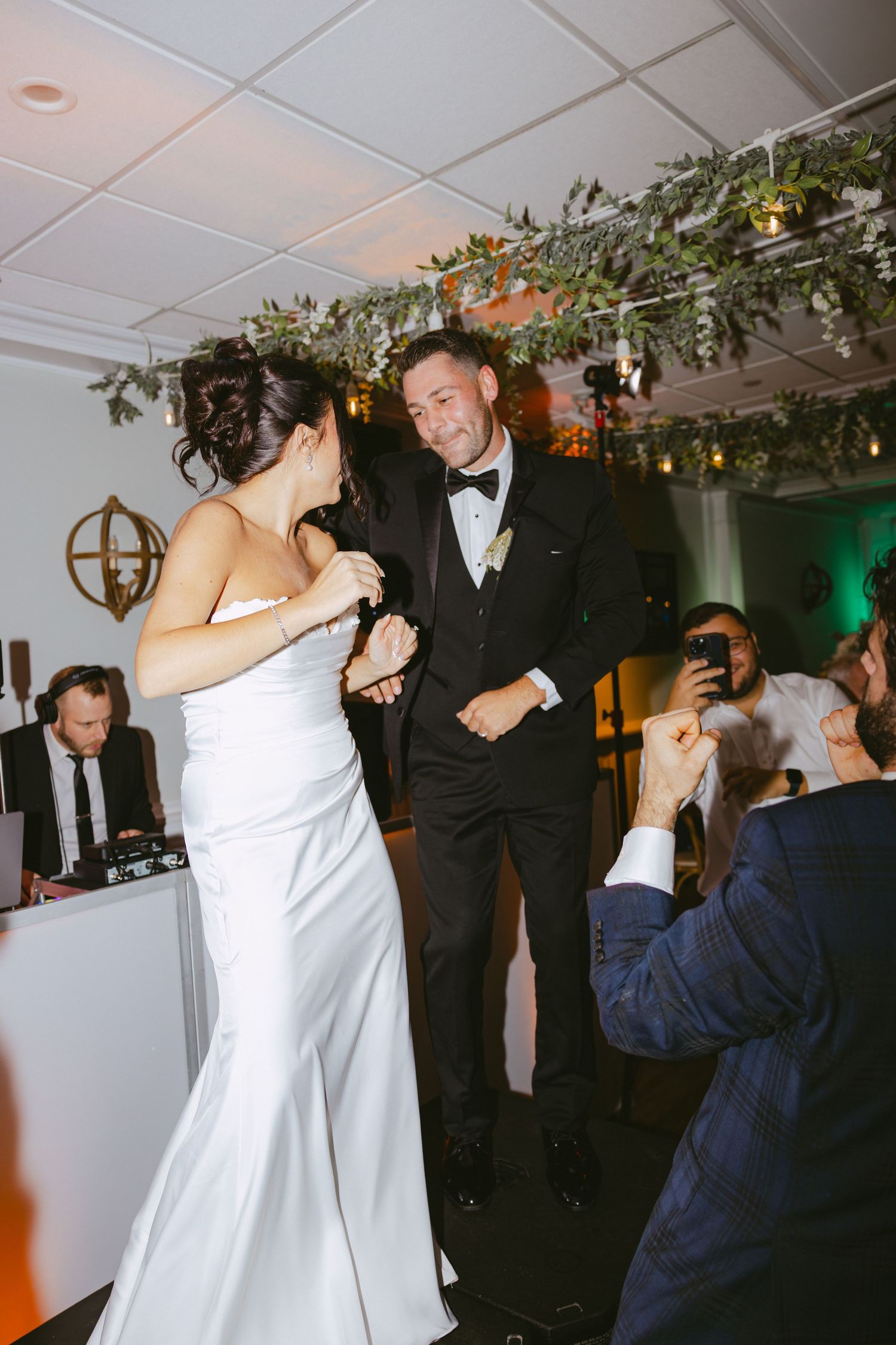 Bride and groom share a joyful first dance at their wedding reception with greenery overhead.