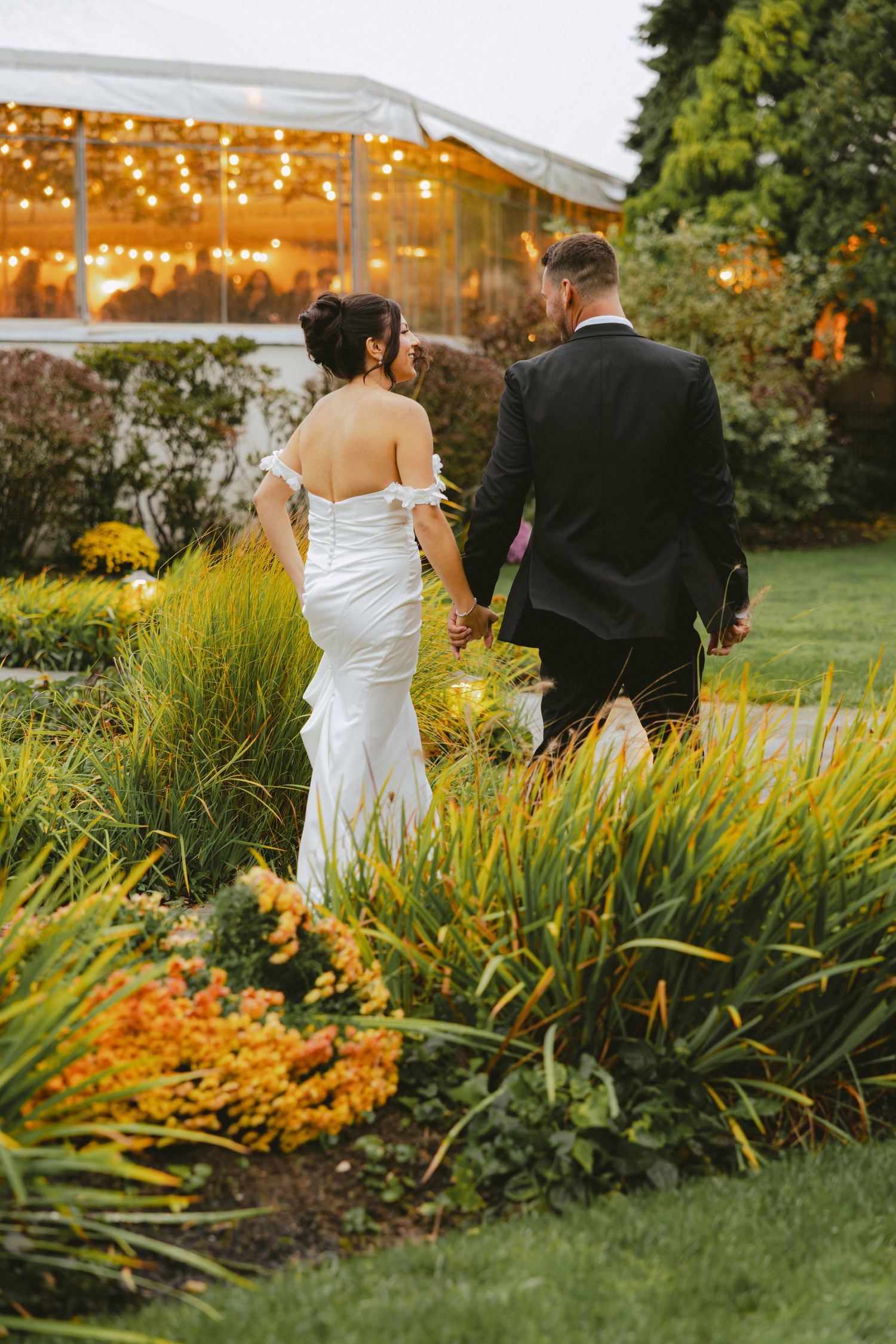 Bride and groom walk hand in hand toward a glowing tent reception venue at dusk.