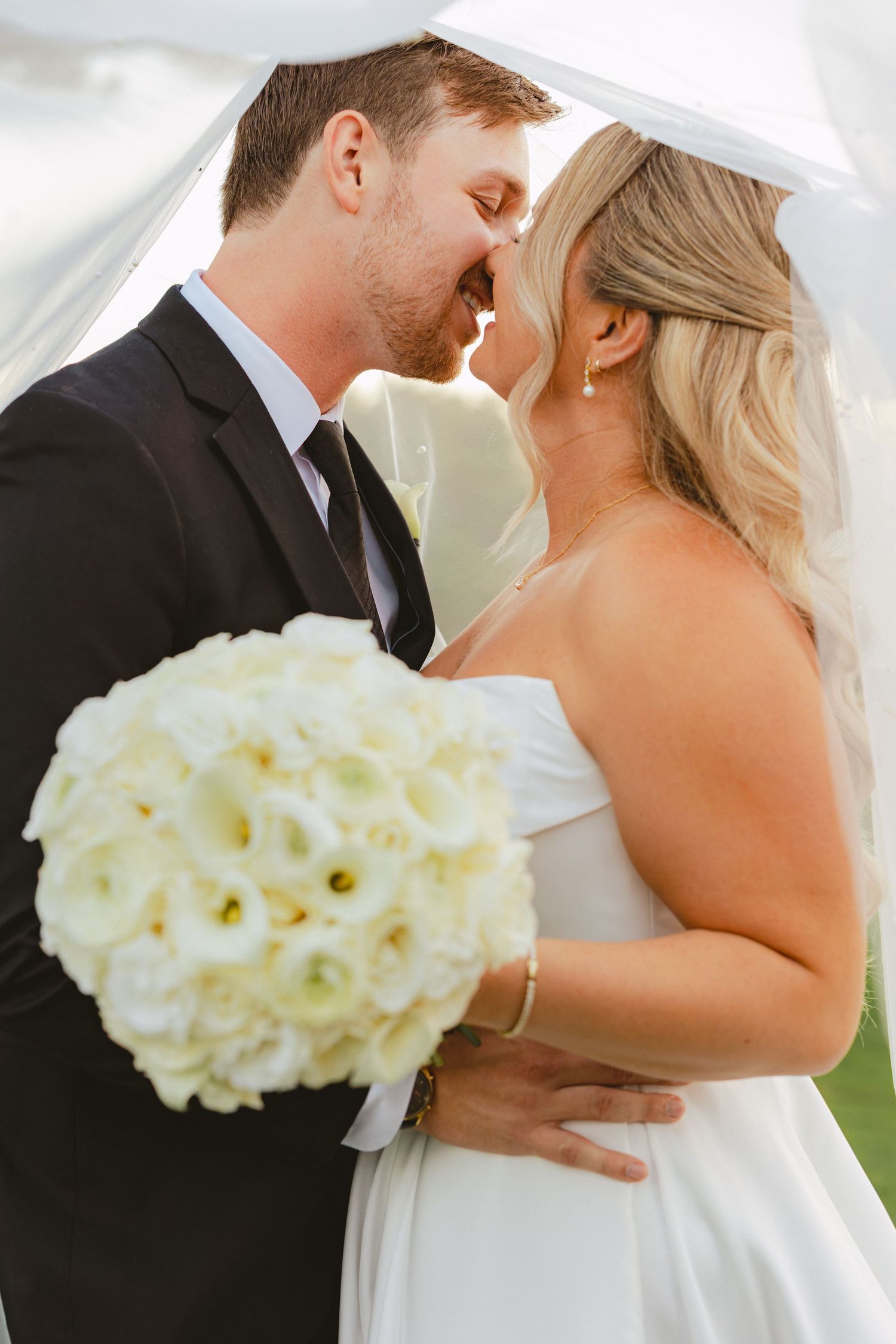 Bride and groom share a romantic almost-kiss under a veil, bride holding a white calla lily bouquet.