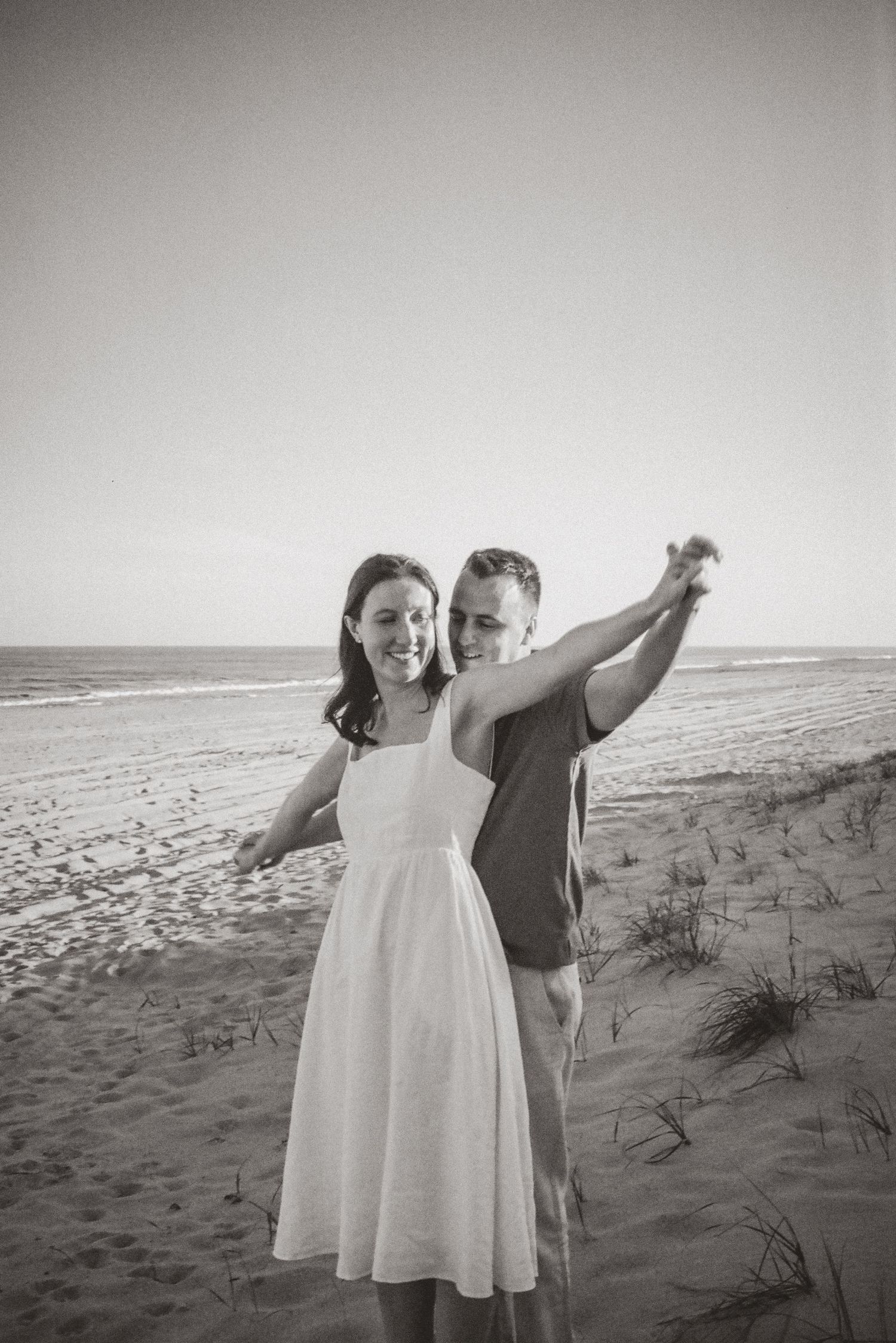 Black and white photo of a joyful couple posing on a sandy beach with arms outstretched near dunes.
