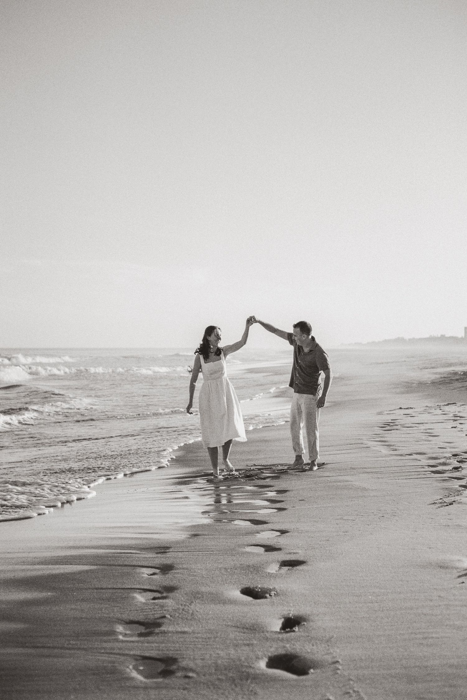 Black and white photo of couple twirling and holding hands along beach shoreline with footprints.
