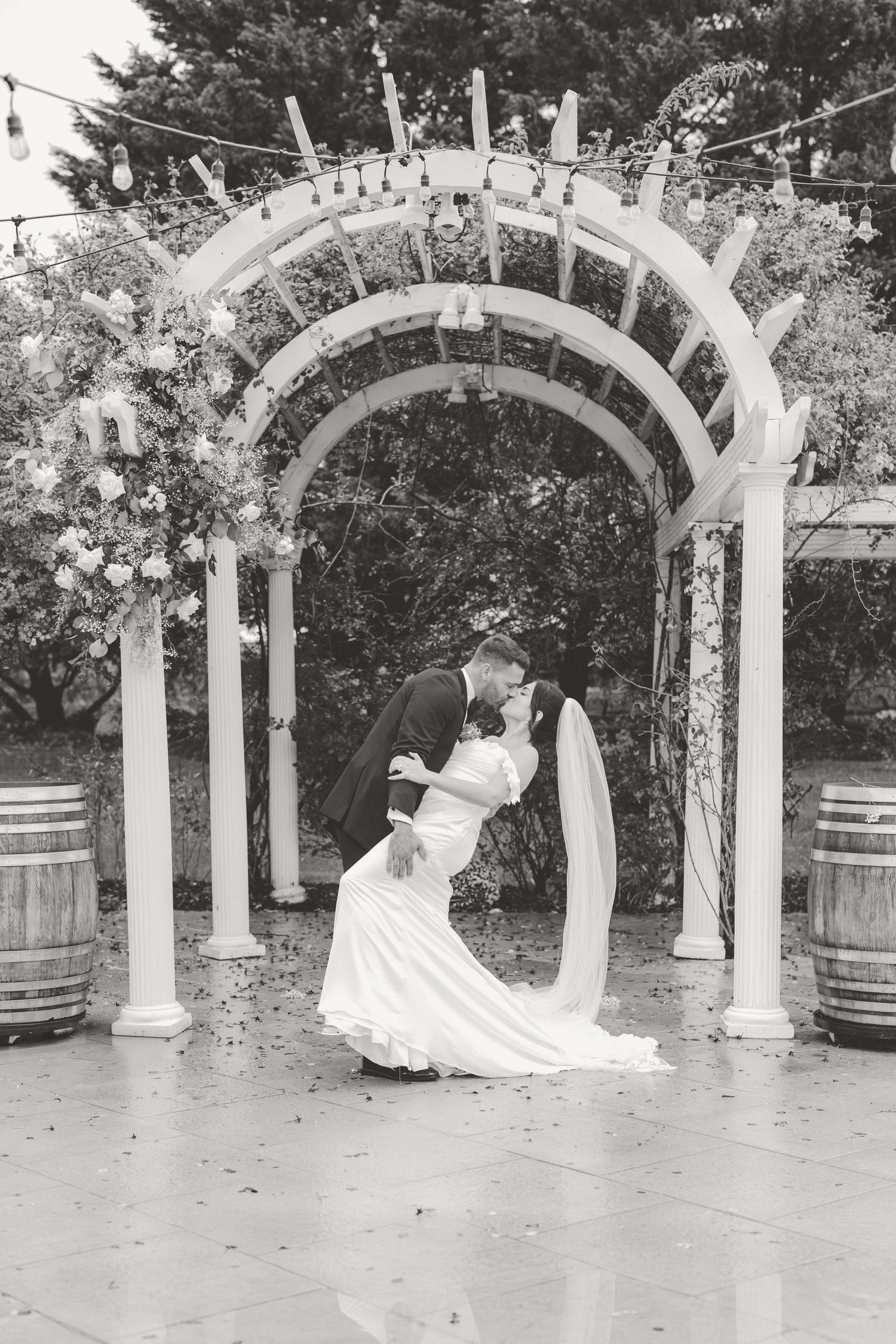 Bride and groom share a romantic dip kiss under a floral arch with string lights outdoors.