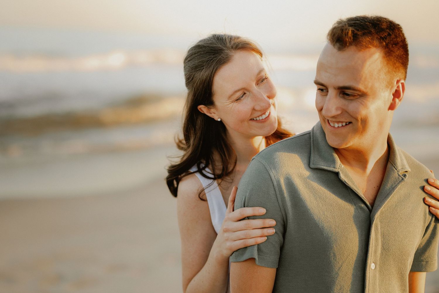 Smiling couple embracing closely at golden hour beach with soft ocean waves in background.