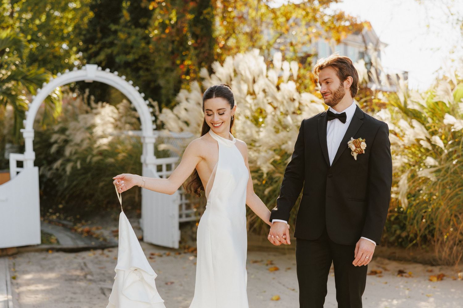 Bride in halter gown and groom in black tuxedo walk hand in hand past white garden arch.
