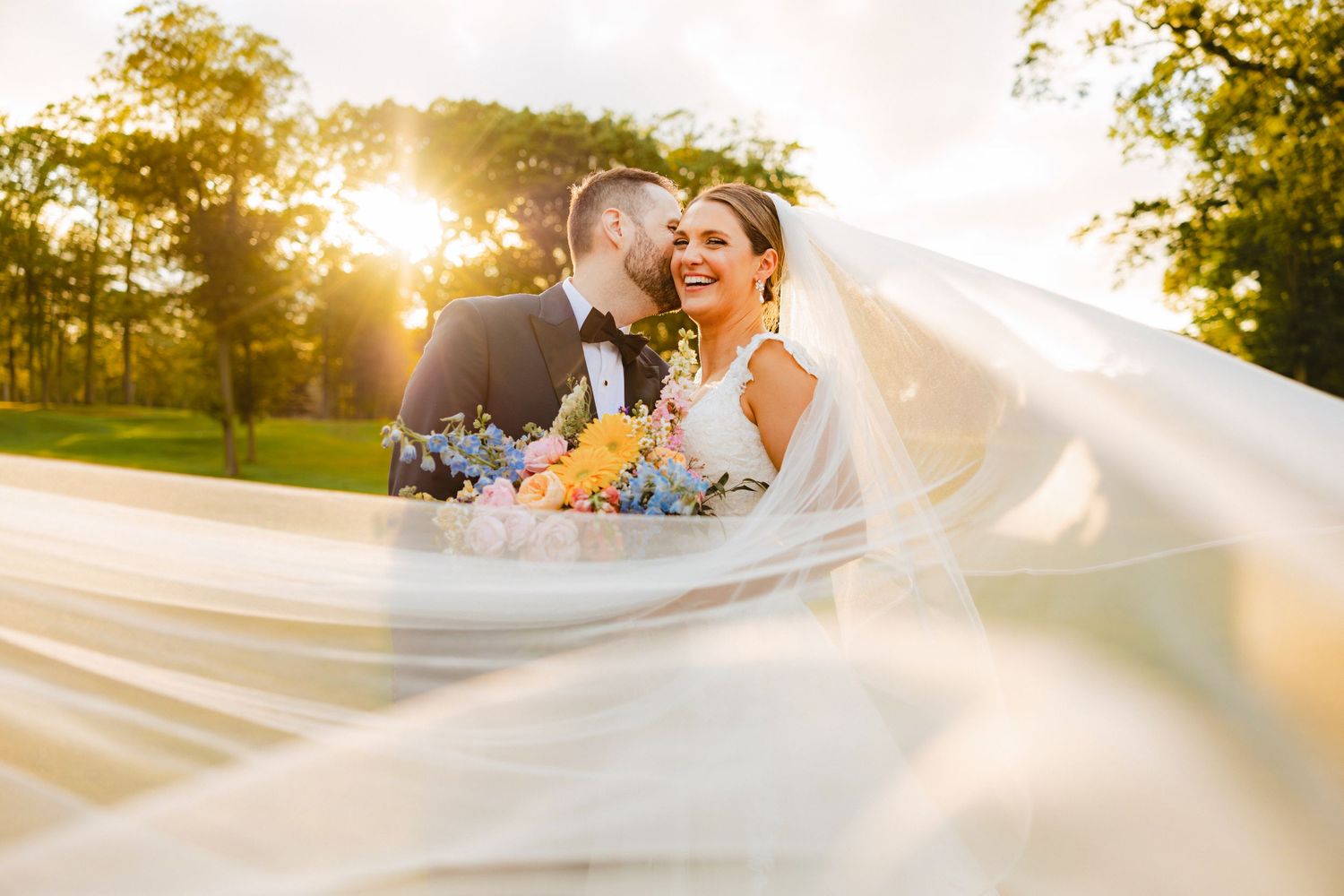 Bride smiles as veil billows in wind while groom kisses her cheek at golden sunset outdoors.