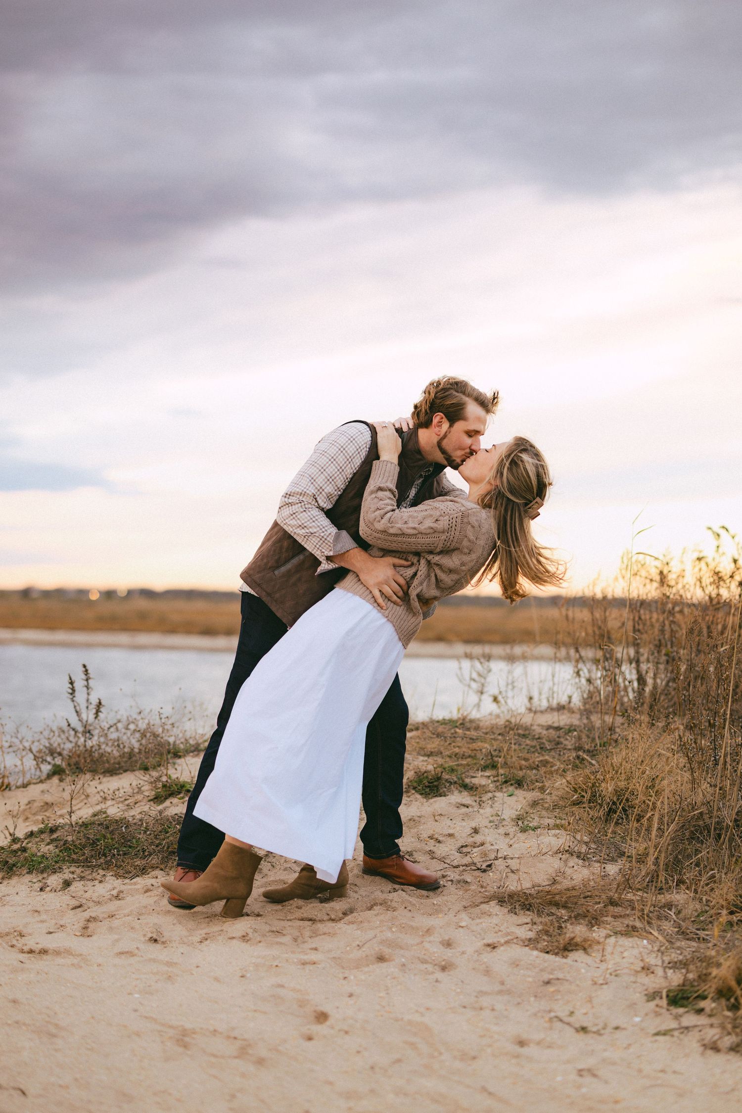 Romantic couple shares a dip kiss on sandy shore at golden hour with tall grass and calm water behind them.