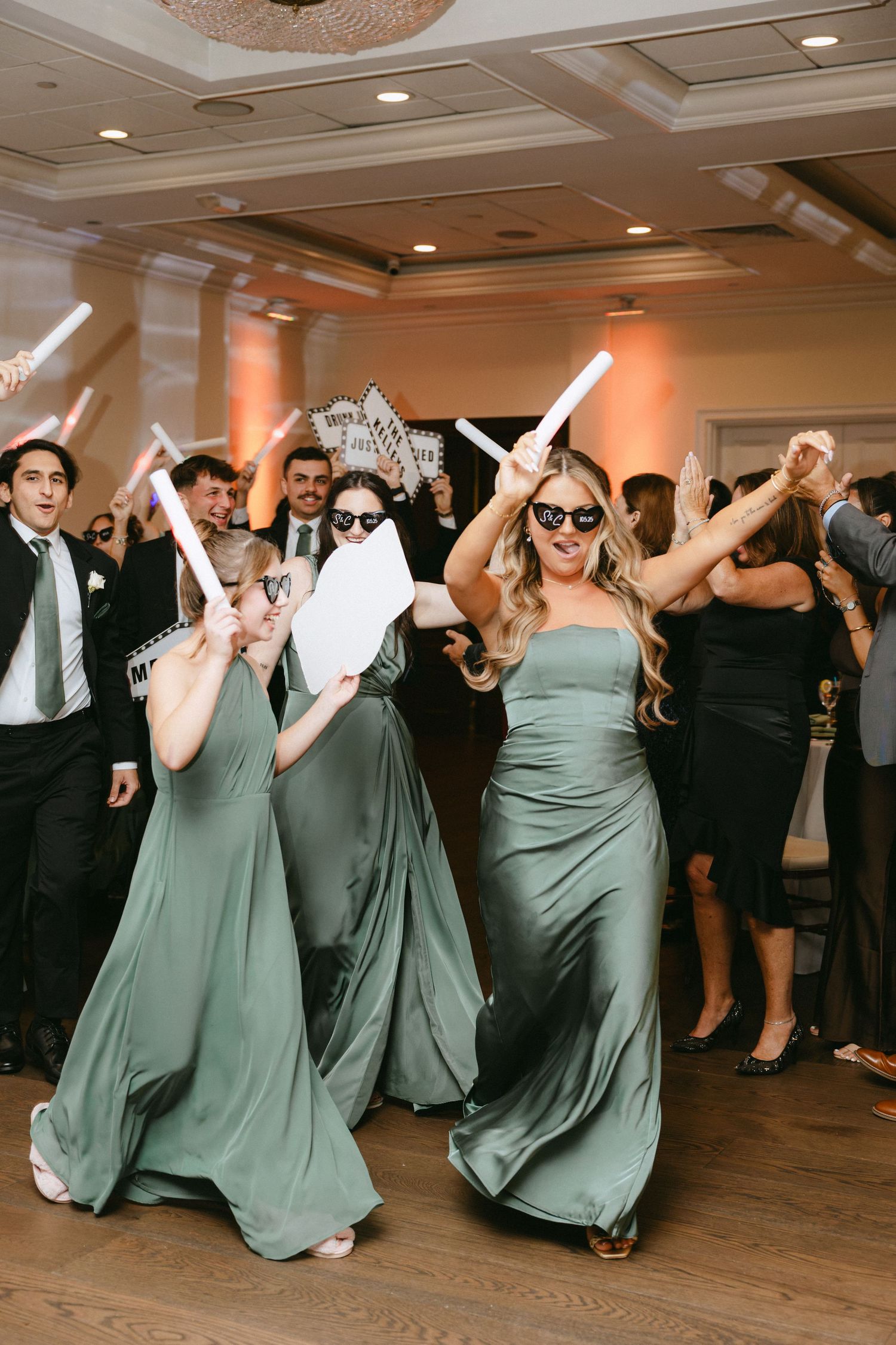 Bridesmaids in sage green gowns dance with glow sticks and sunglasses at lively wedding reception.