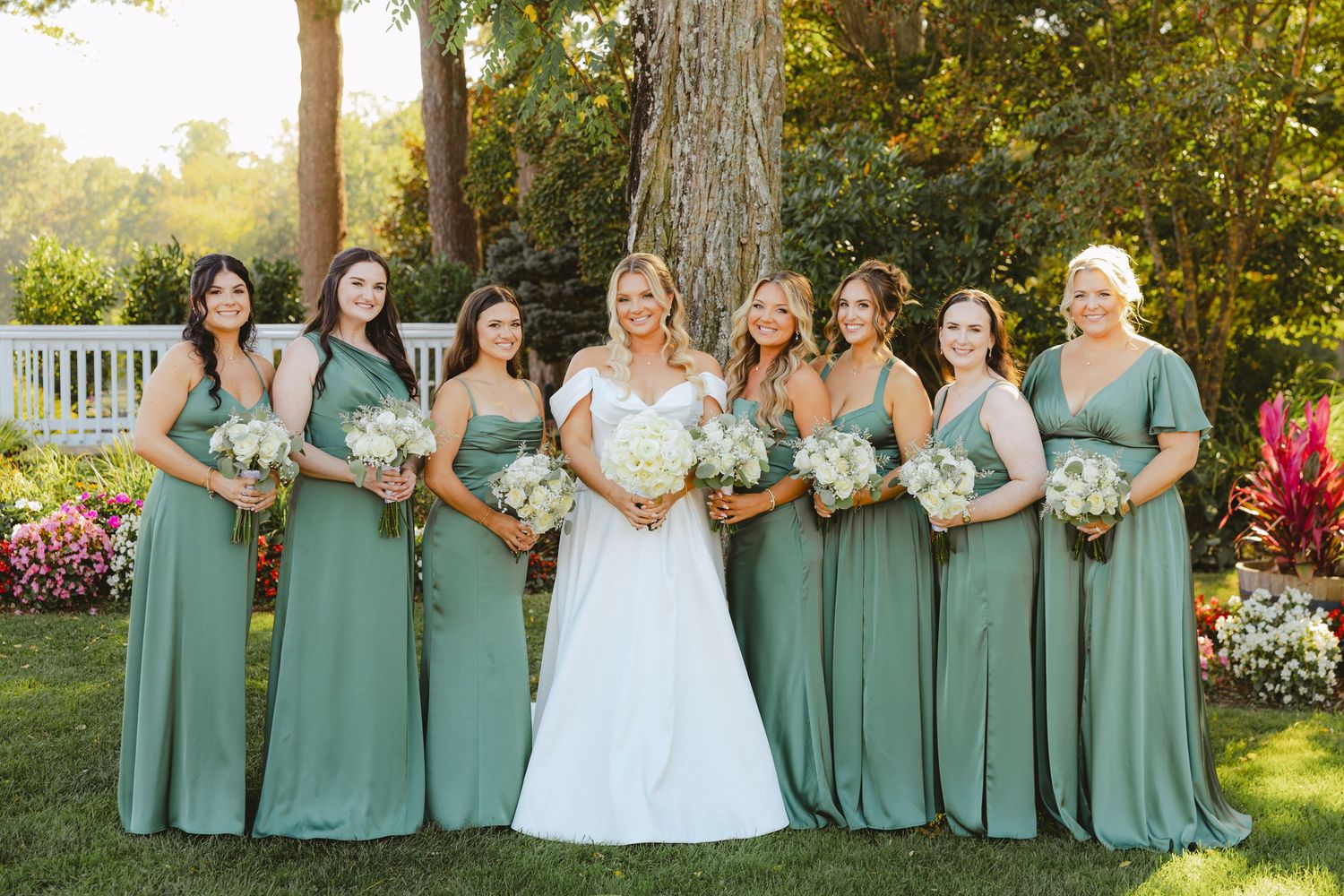 Bride in white gown poses with six bridesmaids in sage green dresses holding white bouquets.