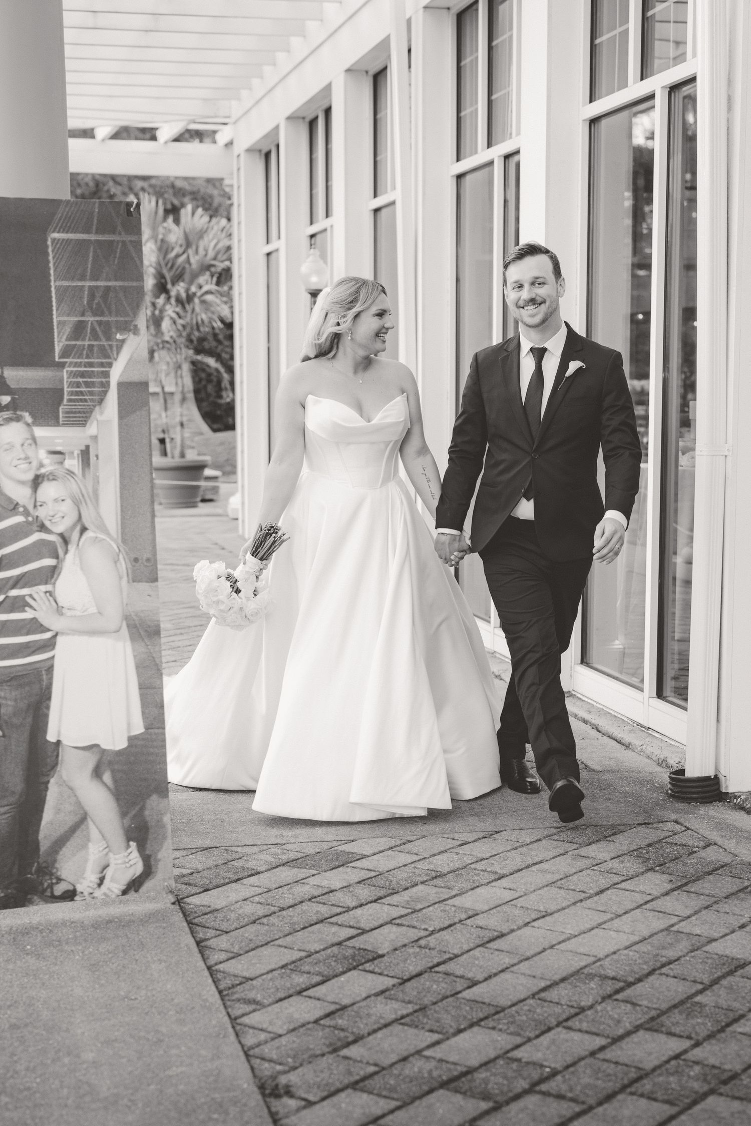 Black and white photo of smiling bride and groom walking hand in hand outside a venue with guests watching.