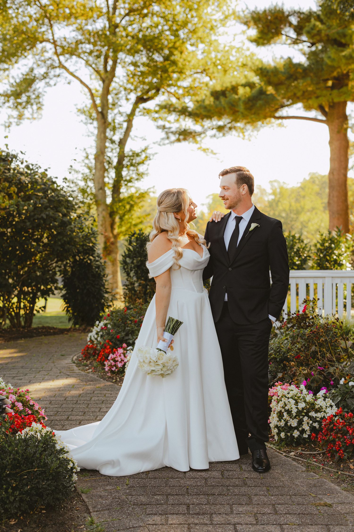 Bride and groom walk together along a brick garden path lined with colorful flowers in soft evening light.