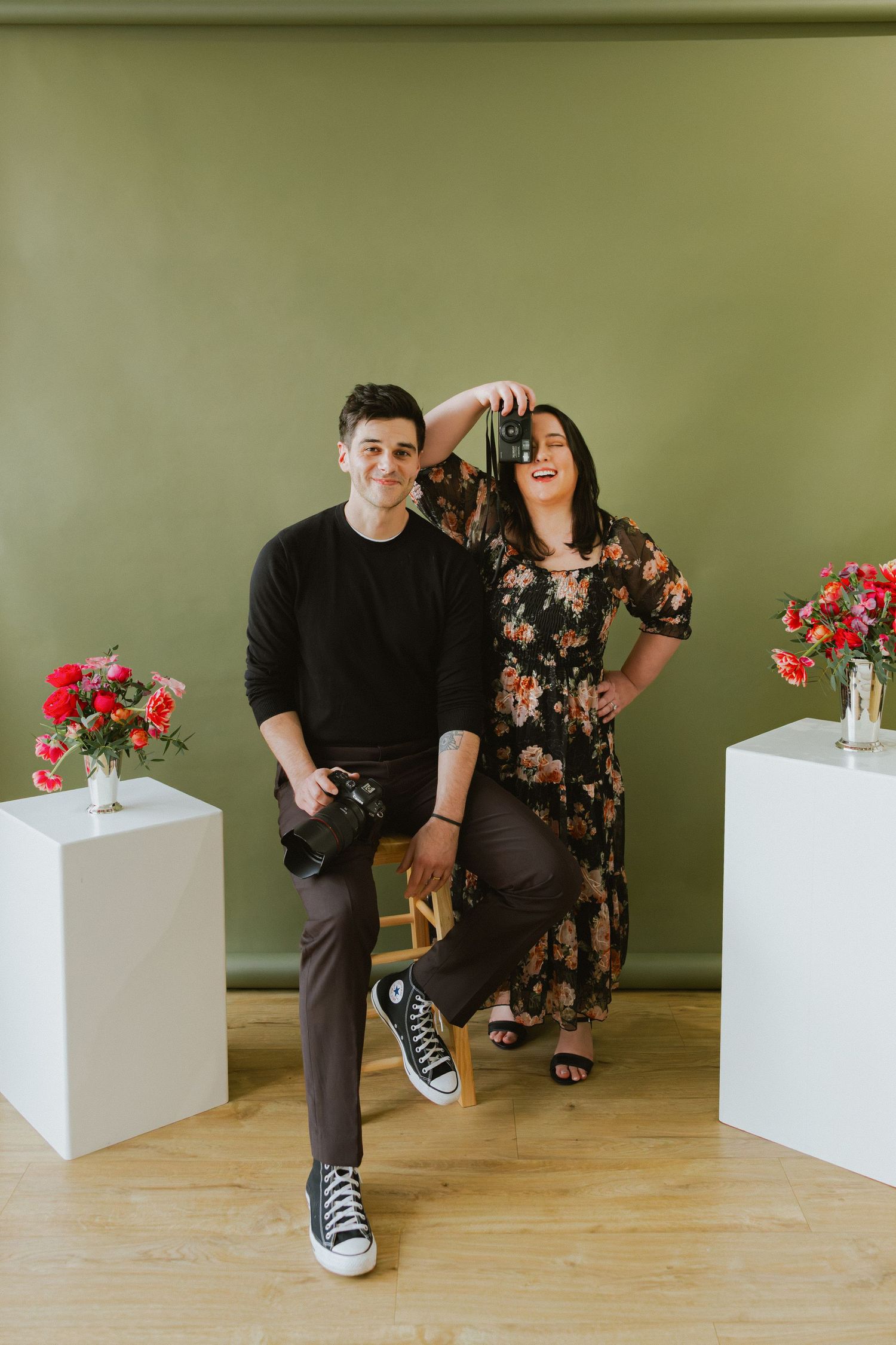 Two wedding photographers pose playfully in a studio with cameras, floral arrangements, and an olive green backdrop.
