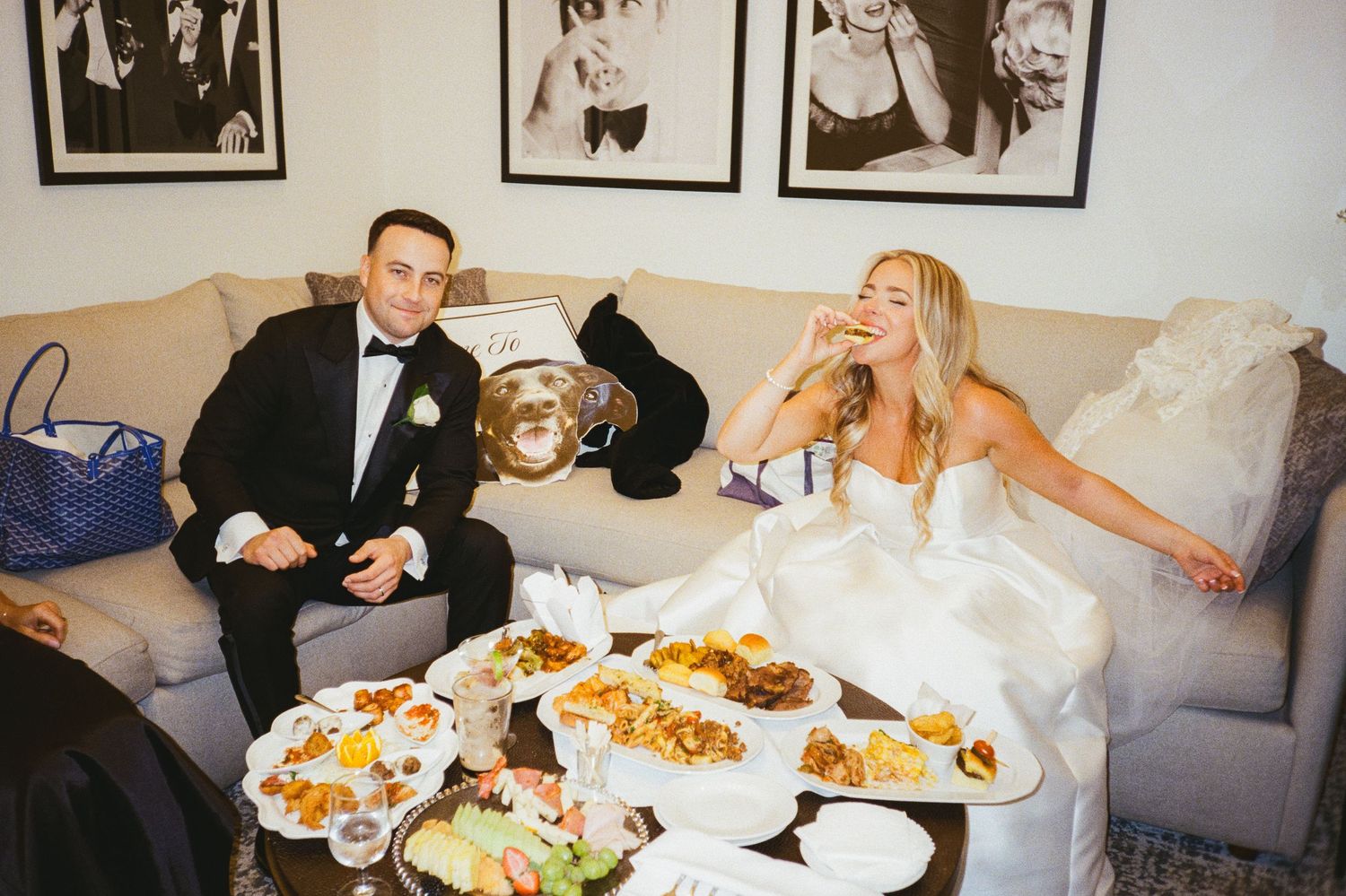 Bride and groom relaxing on a sofa enjoying a late-night snack spread at reception.