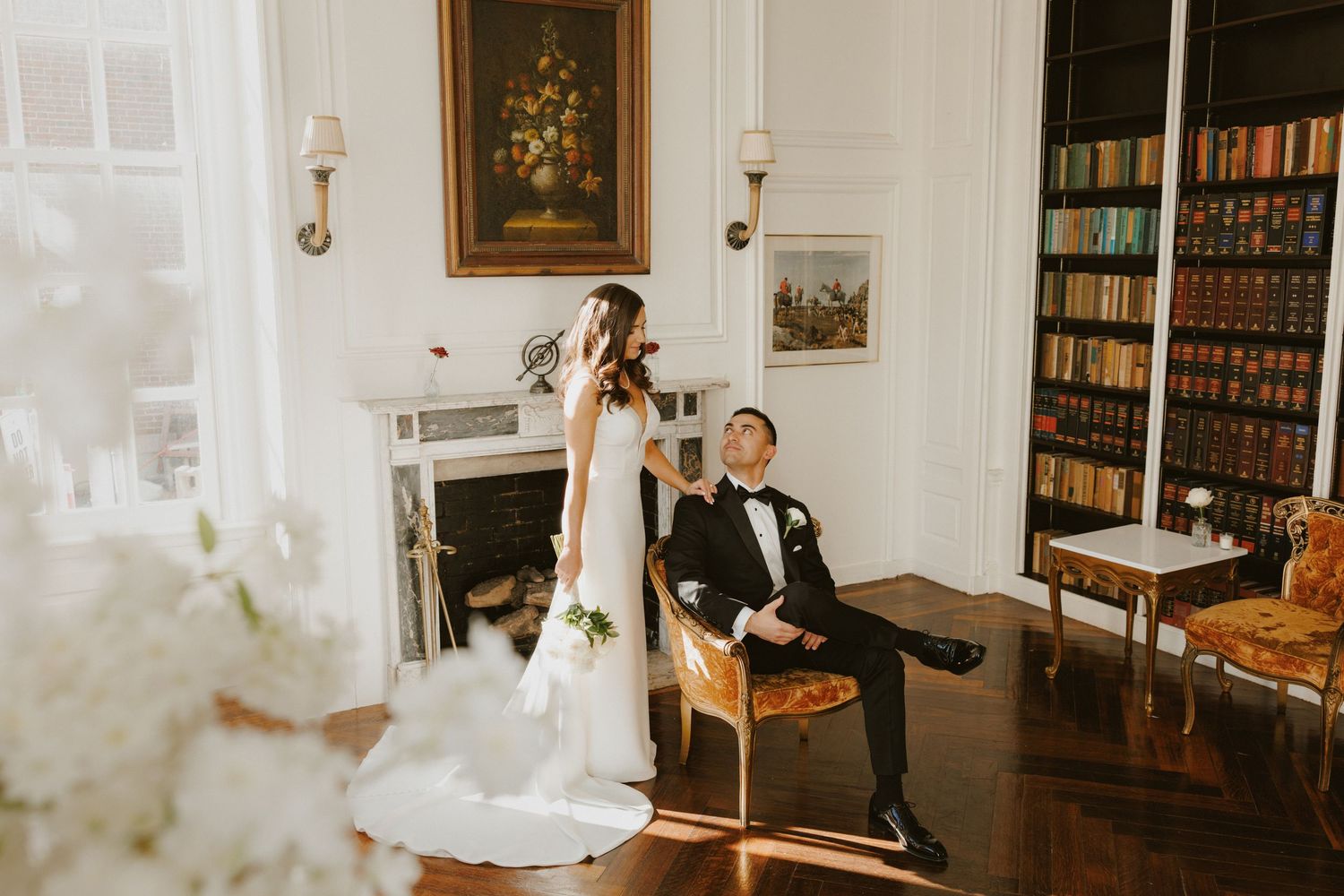Bride in elegant gown stands beside seated groom in sunlit library with fireplace and bookshelves.