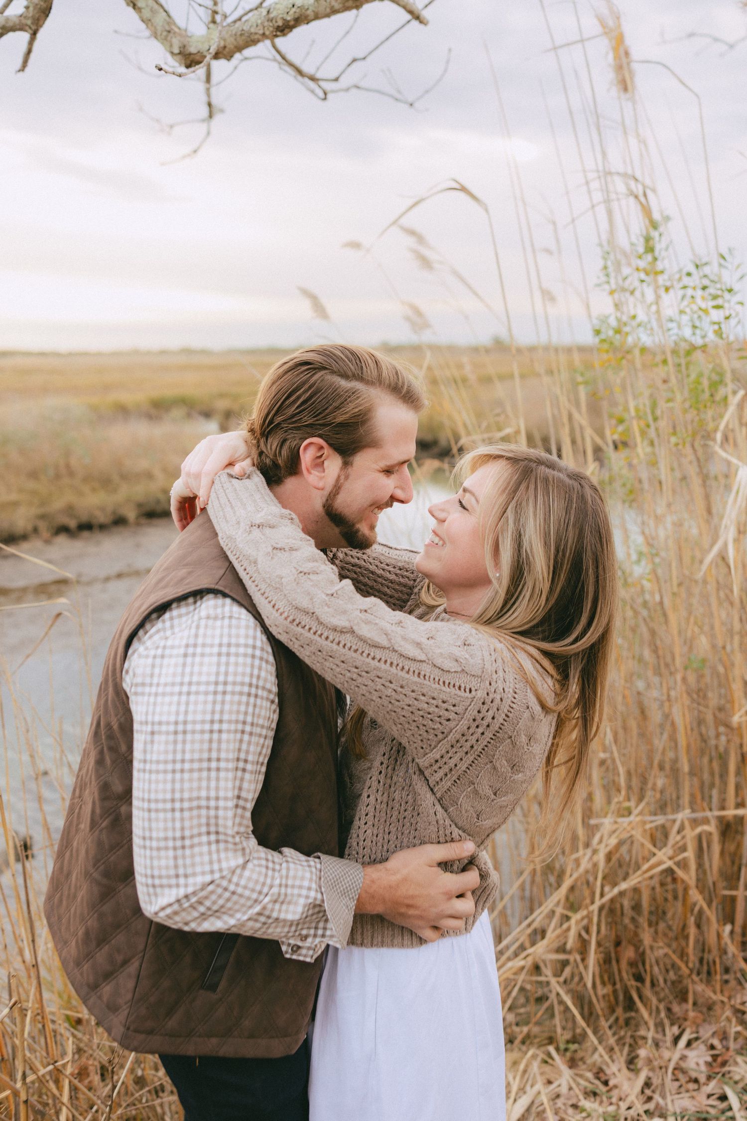 Couple embracing and smiling at each other in a golden marsh landscape under a cloudy sky.