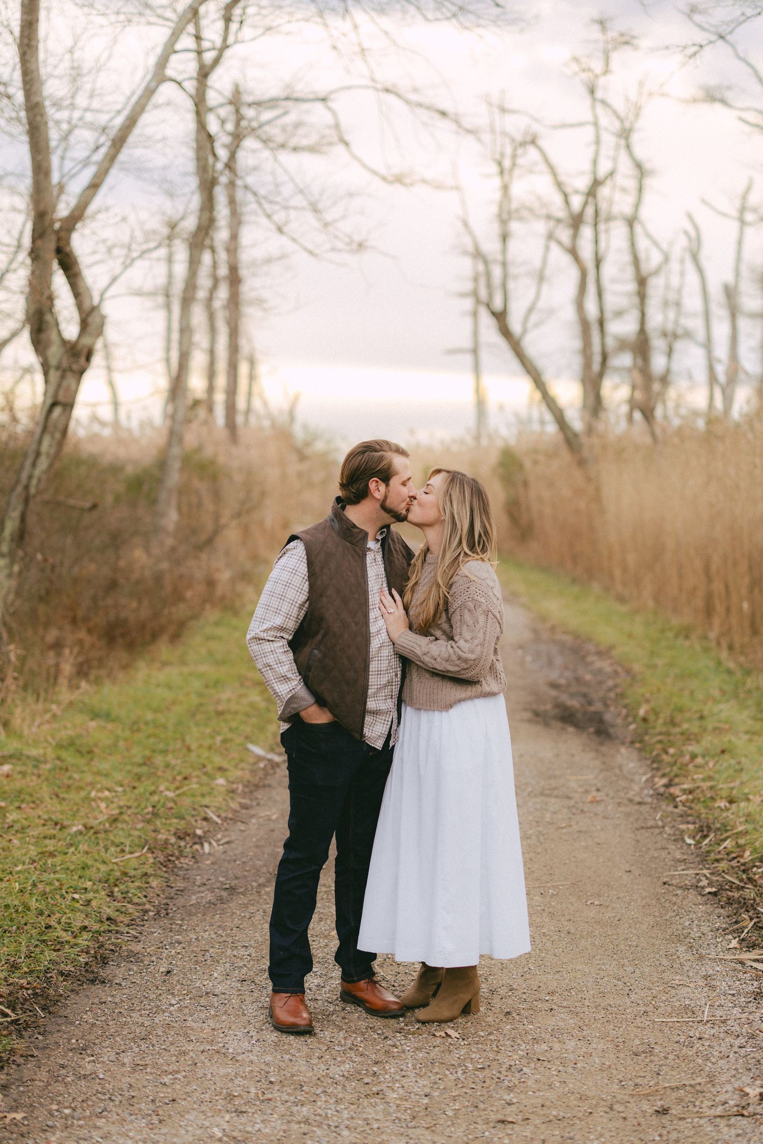 Couple sharing a romantic kiss on a rustic gravel path surrounded by autumn trees and golden grass.