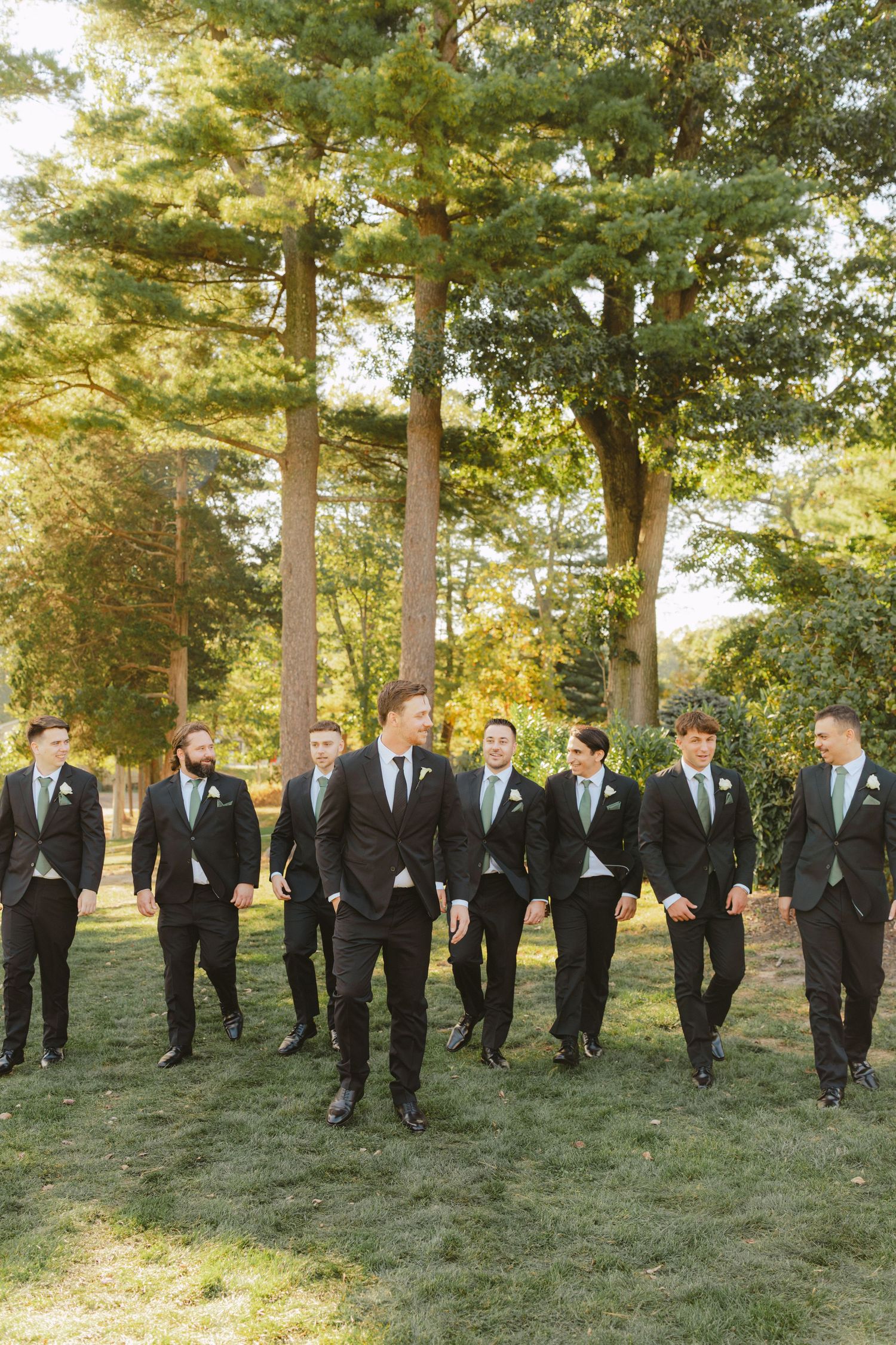 Groom leads groomsmen in black suits with sage green ties walking across sunny outdoor lawn.