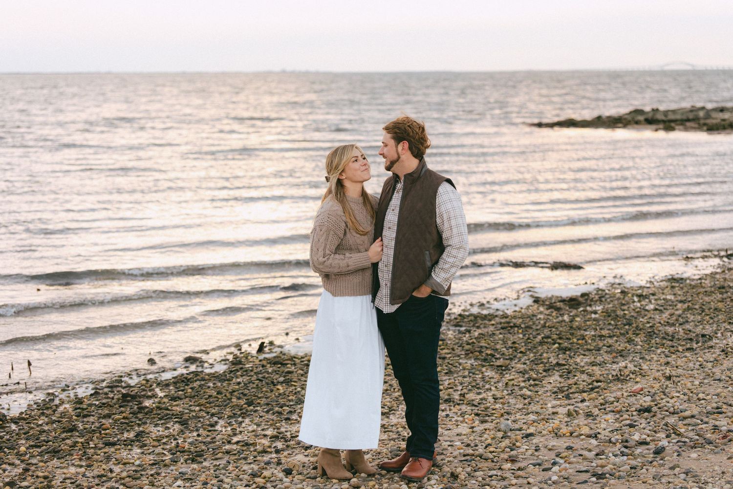 Couple standing close together on a rocky beach shore at golden hour, gazing into each other's eyes.