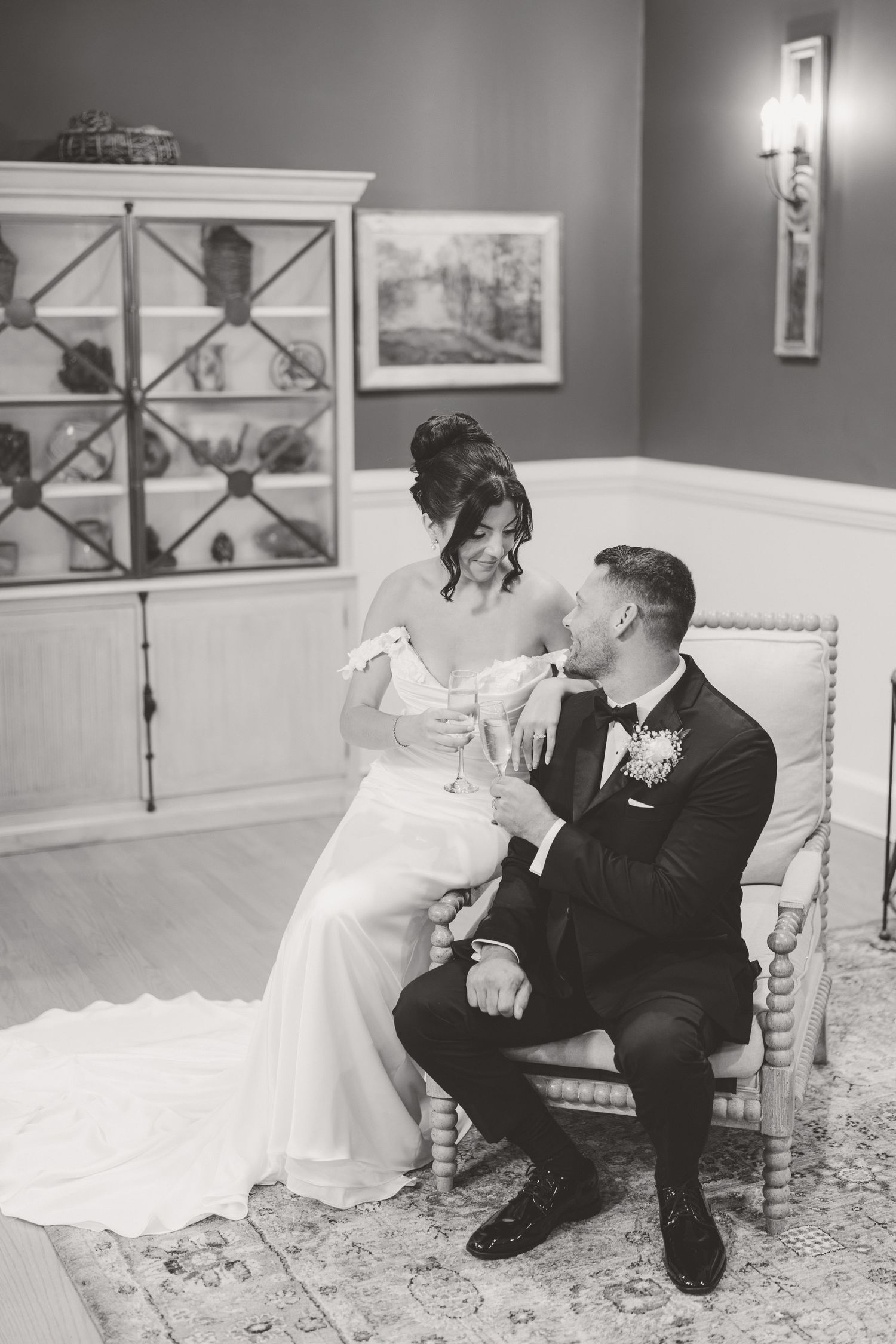 Bride and groom share an intimate moment seated in an elegant chair, holding champagne glasses.