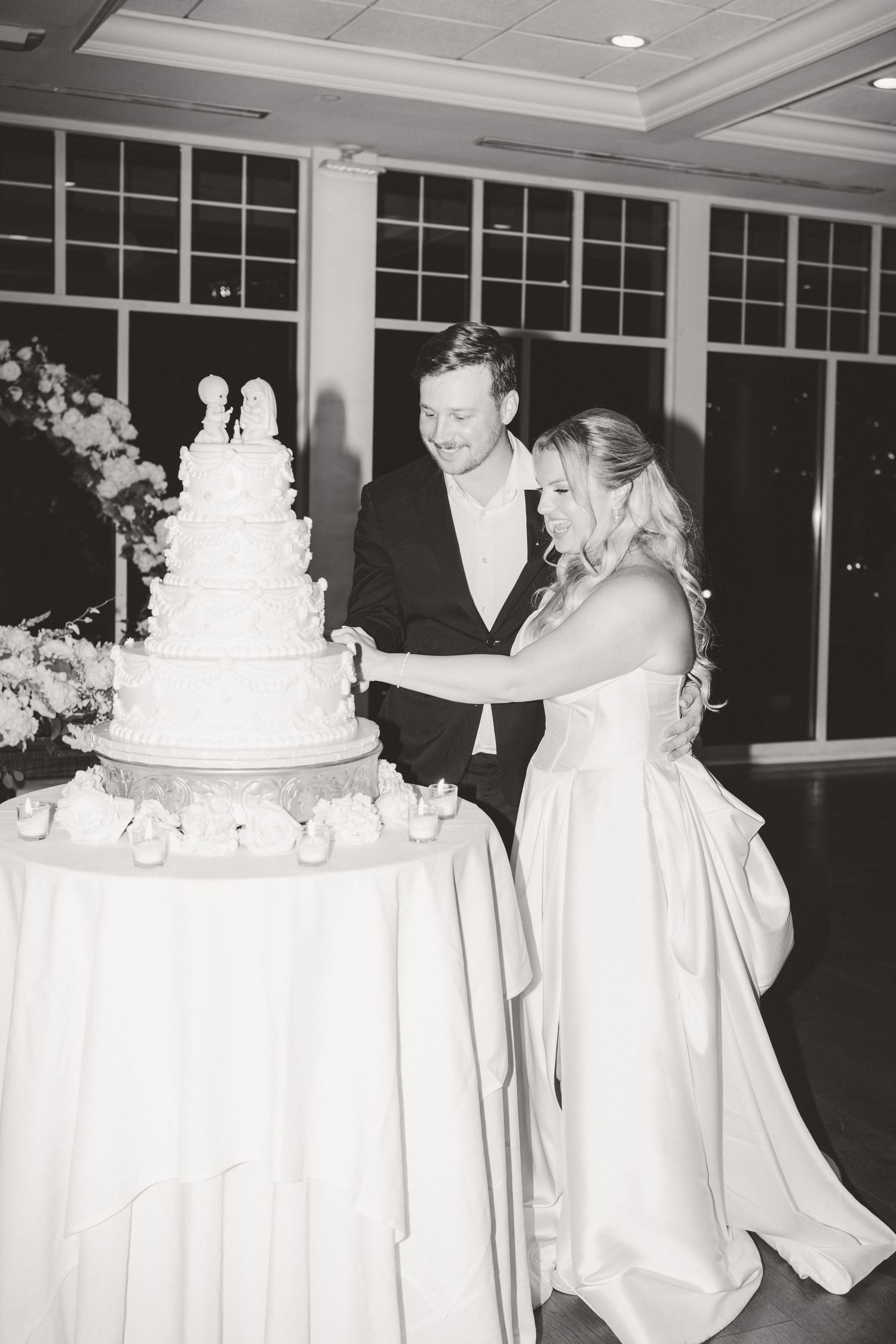 Black and white photo of bride and groom smiling while cutting their tiered wedding cake at the reception.