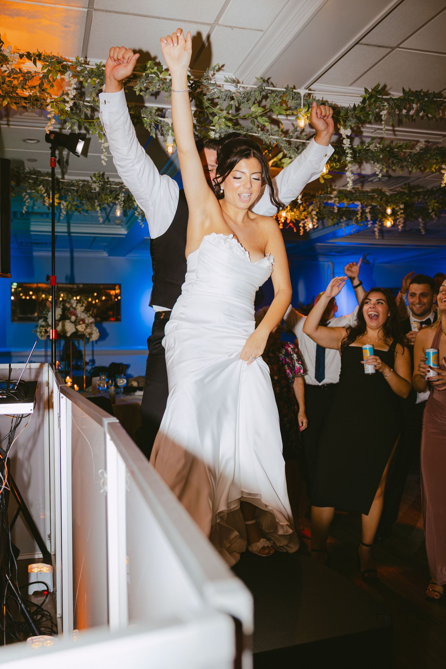 Excited bride raises her arms while dancing with guests celebrating at wedding reception.