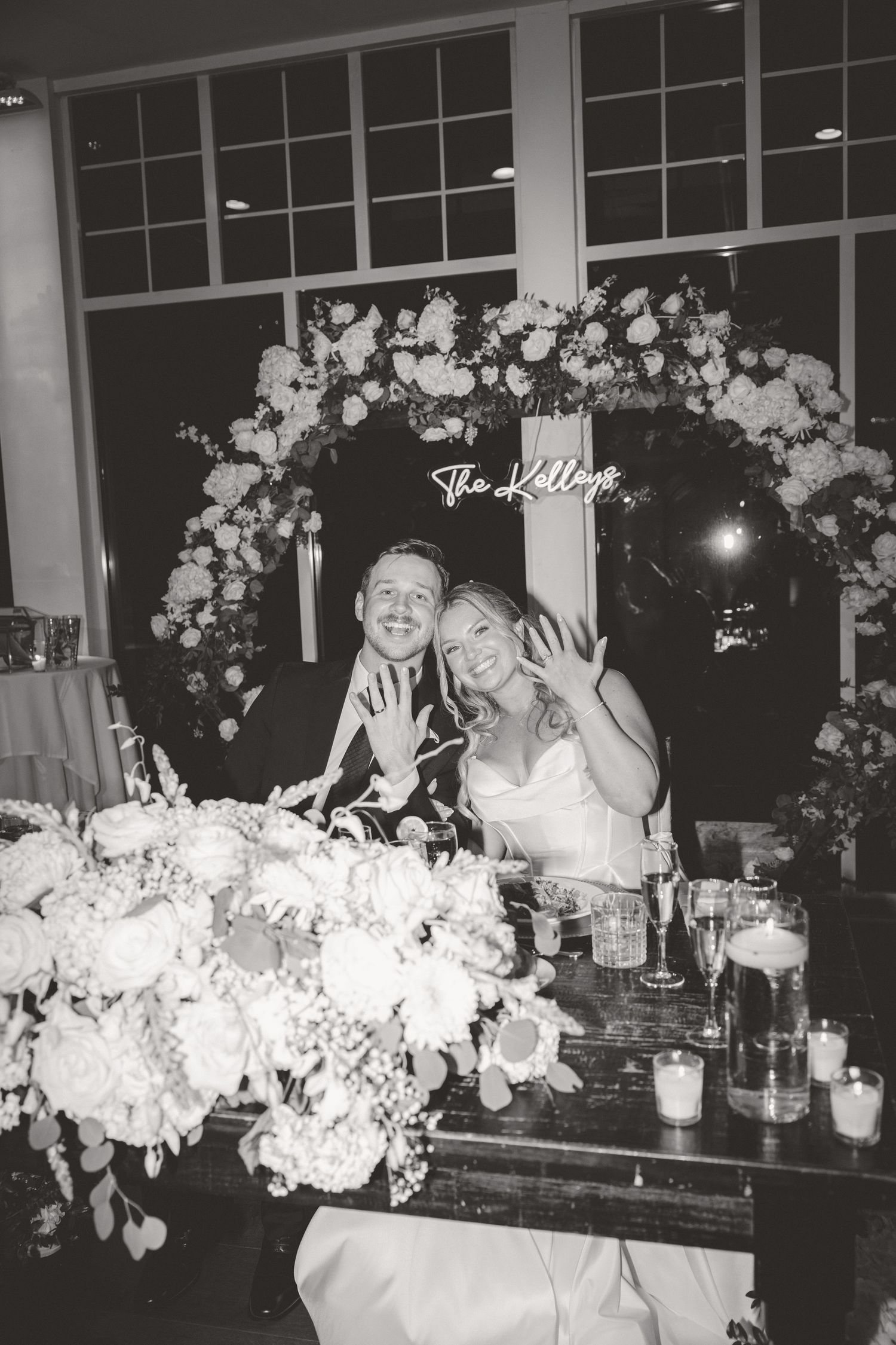 Newlyweds sitting at sweetheart table showing off rings, floral arch with neon sign reading The Kelleys behind them.