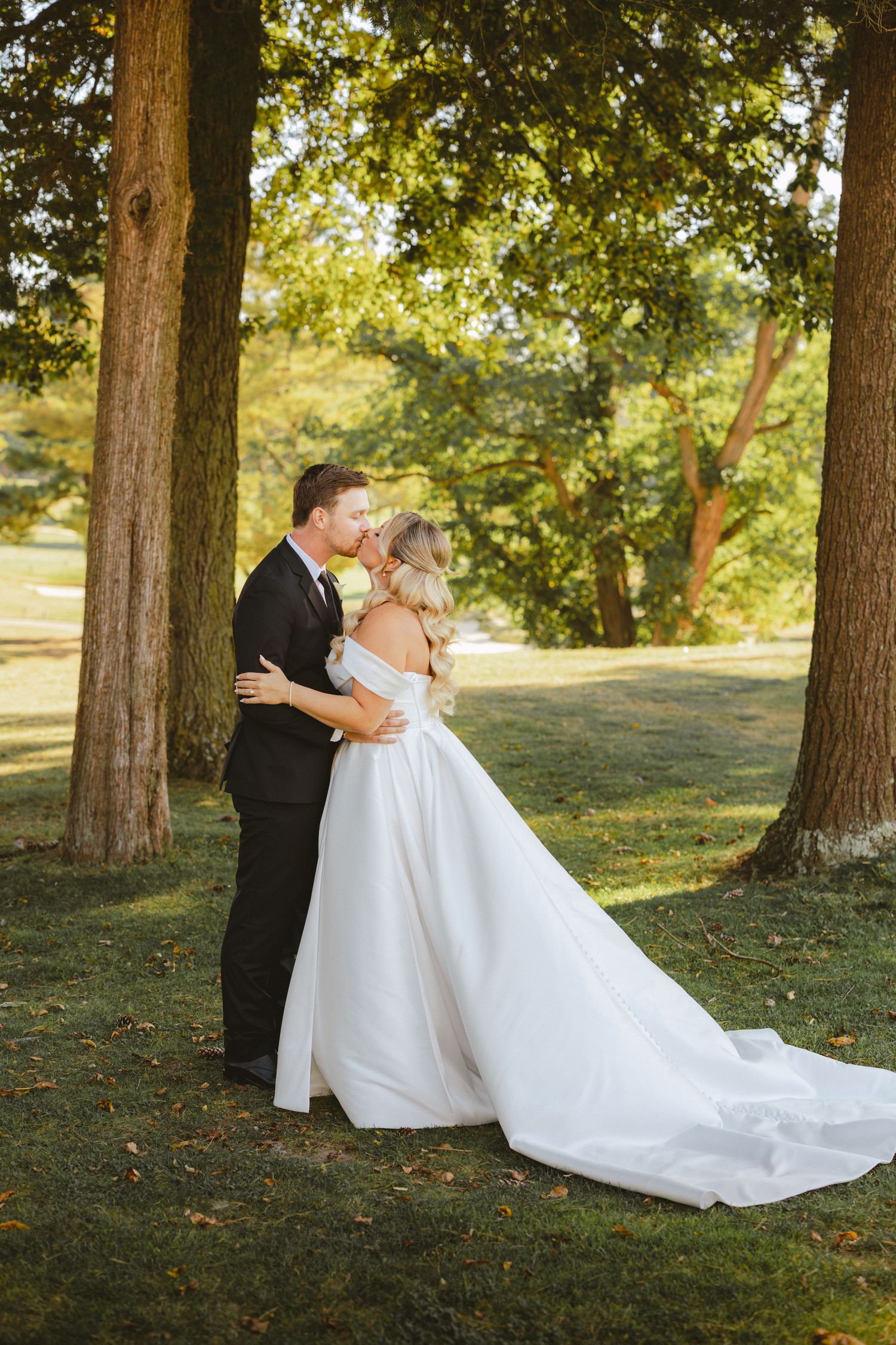 Bride and groom share a kiss beneath tall trees on a green lawn in warm golden afternoon sunlight.
