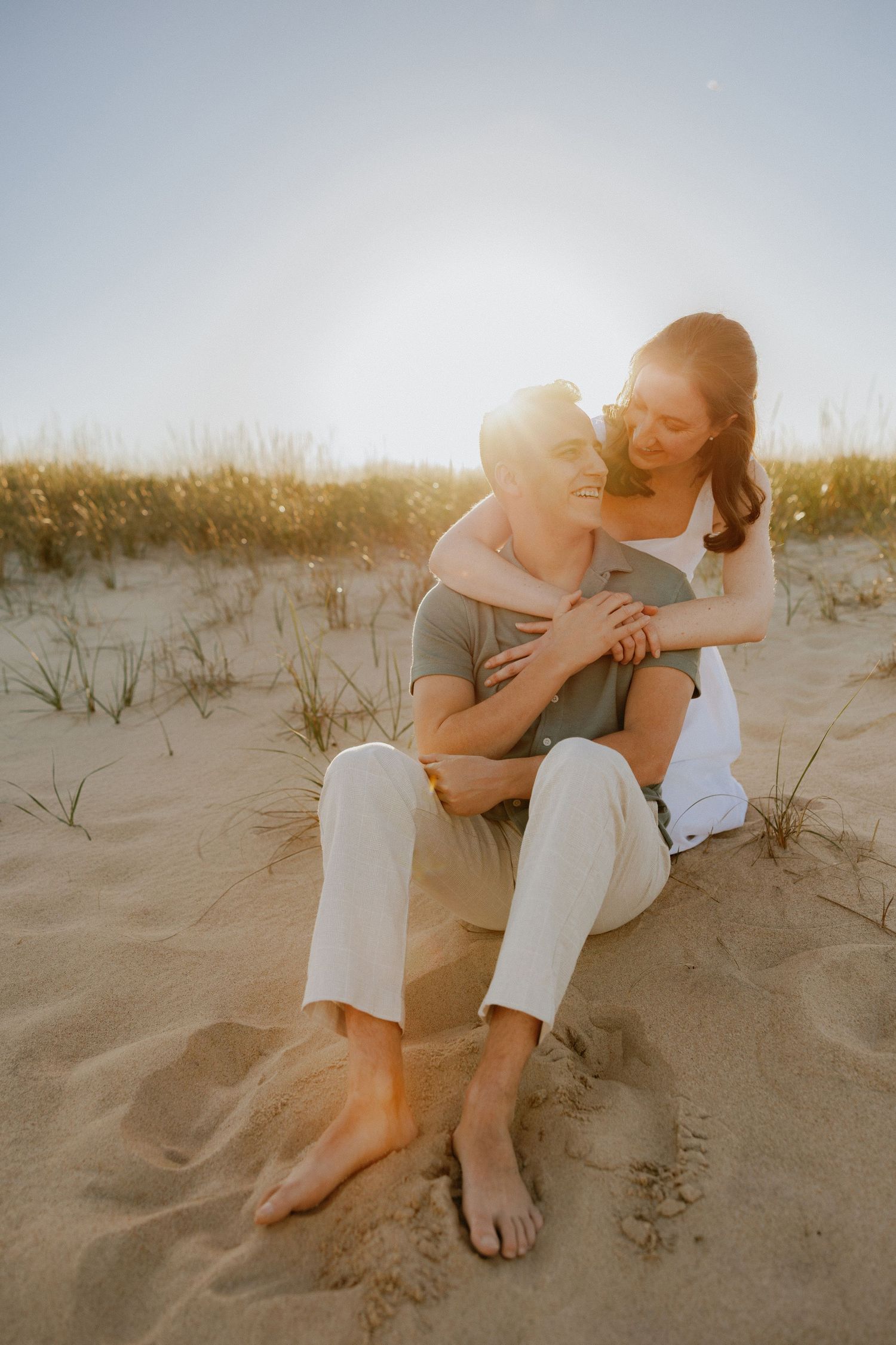 Couple sitting on beach sand dunes at sunset, woman embracing man from behind, smiling joyfully.
