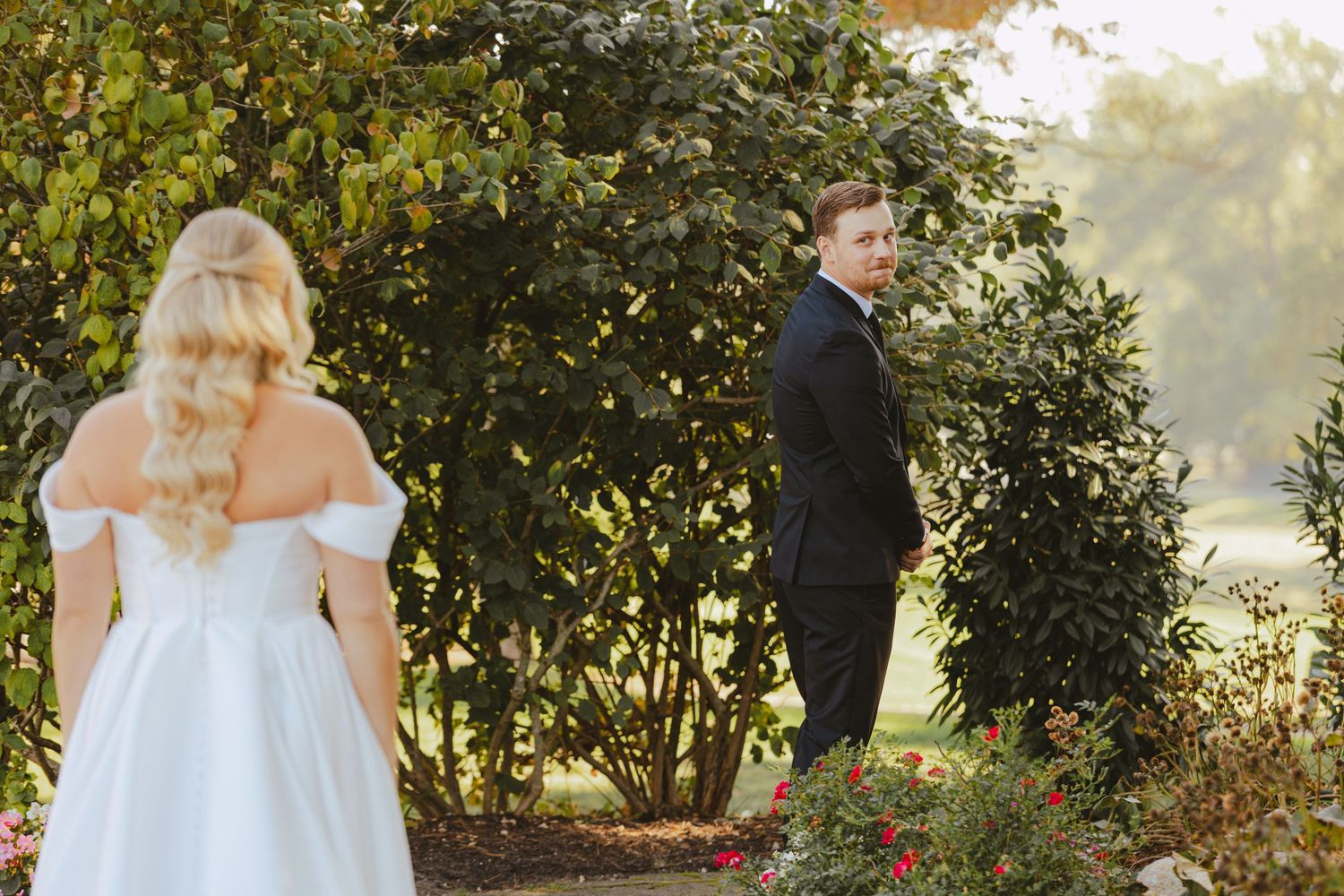 Groom in black tuxedo waits for his first look as bride in white gown approaches garden path.