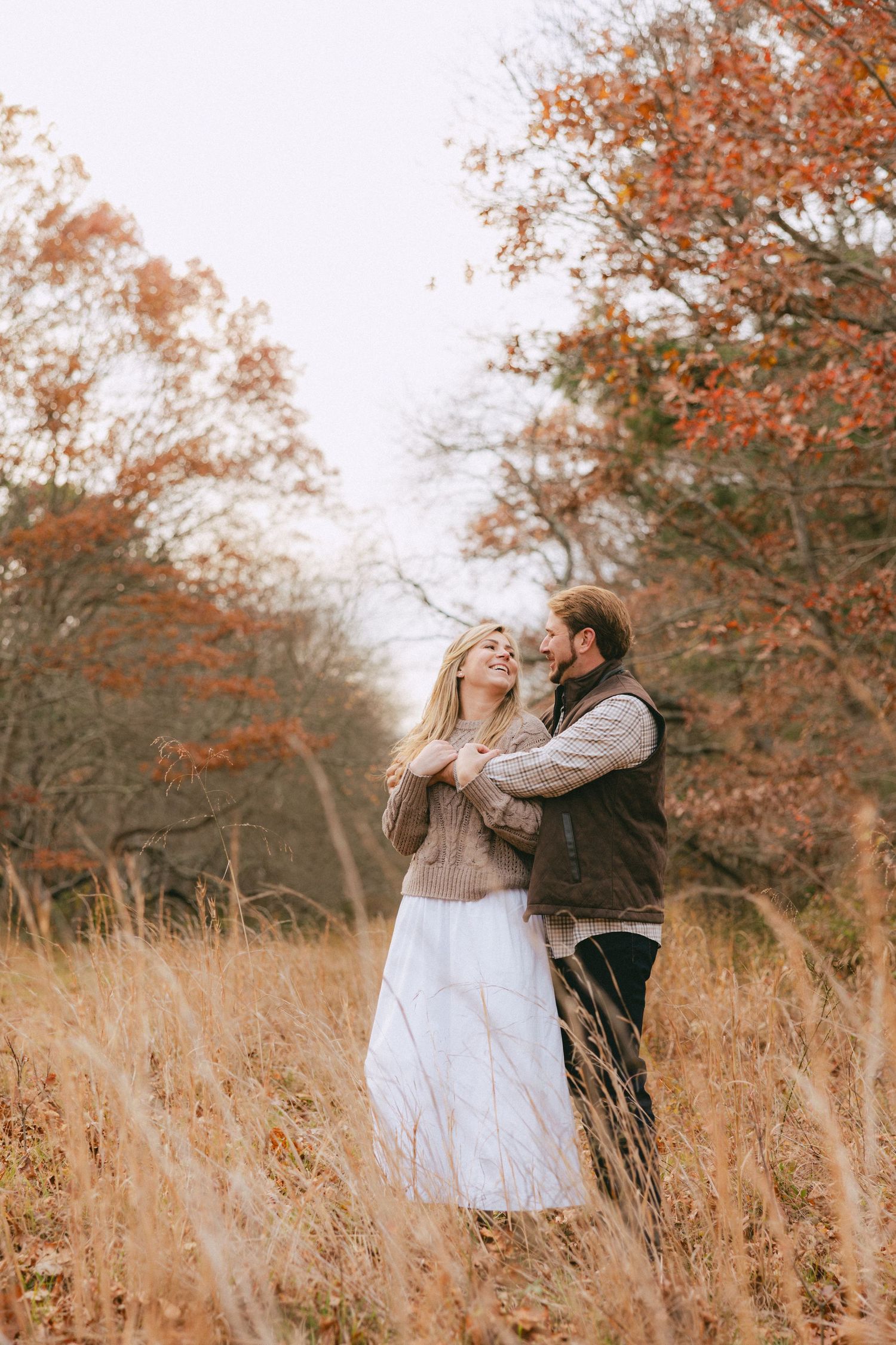Romantic couple embracing in a tall golden grass field beneath stunning red and orange autumn trees.