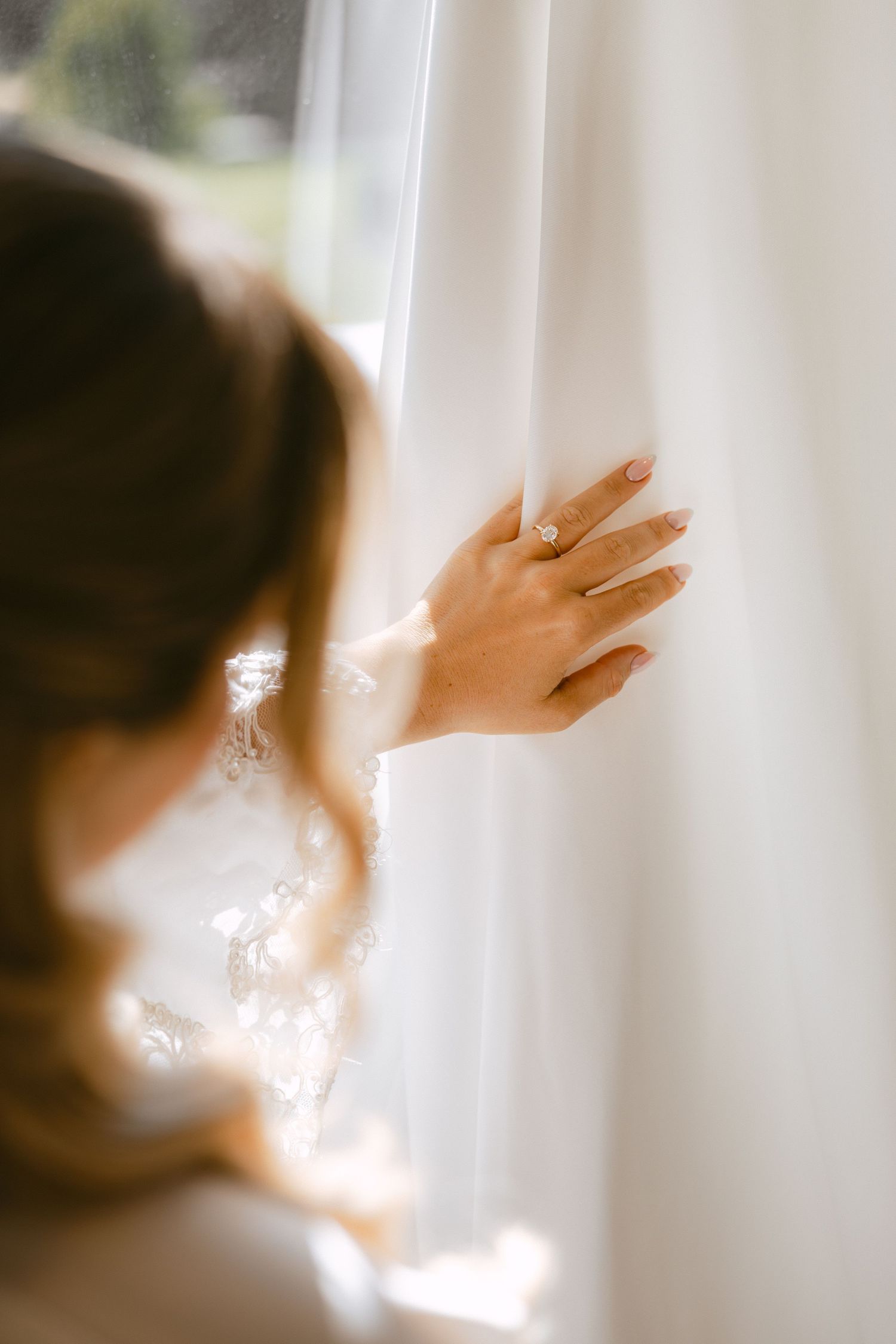 Close-up of bride's hand with engagement ring gently touching white wedding dress near a sunlit window.