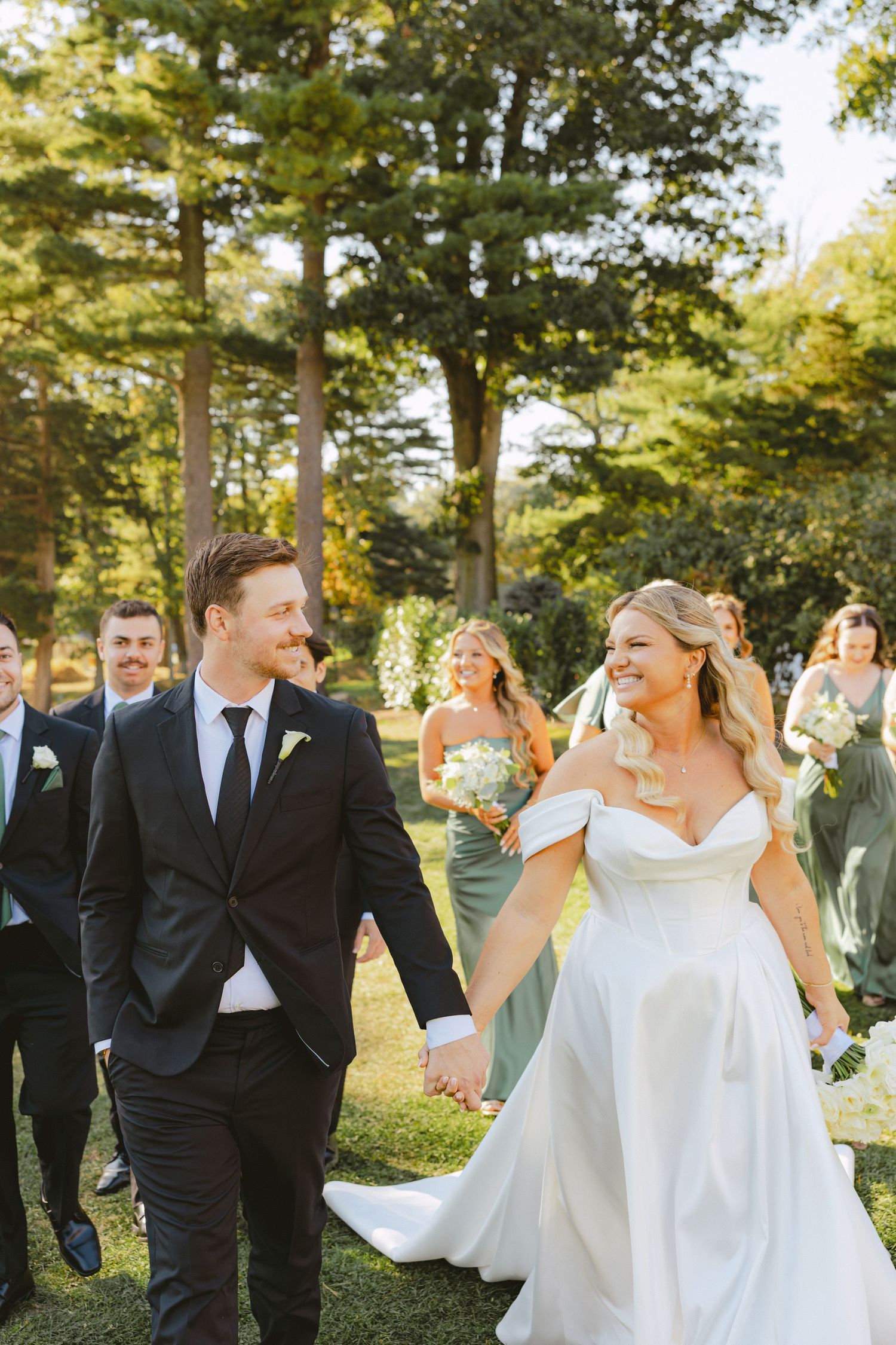 Bride and groom walk hand in hand with wedding party through lush green grounds on their wedding day.