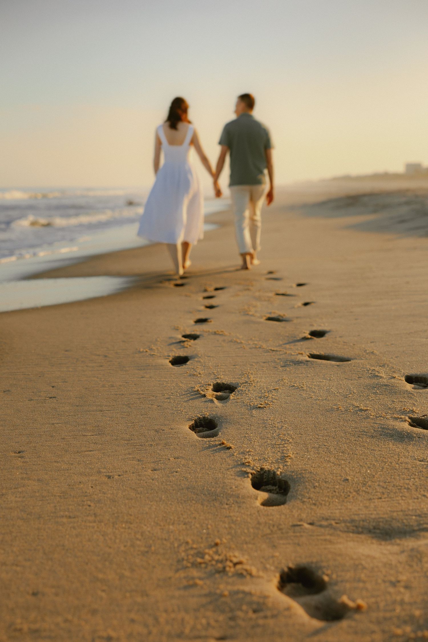 Couple walks hand in hand along sandy beach leaving footprints at golden sunset.