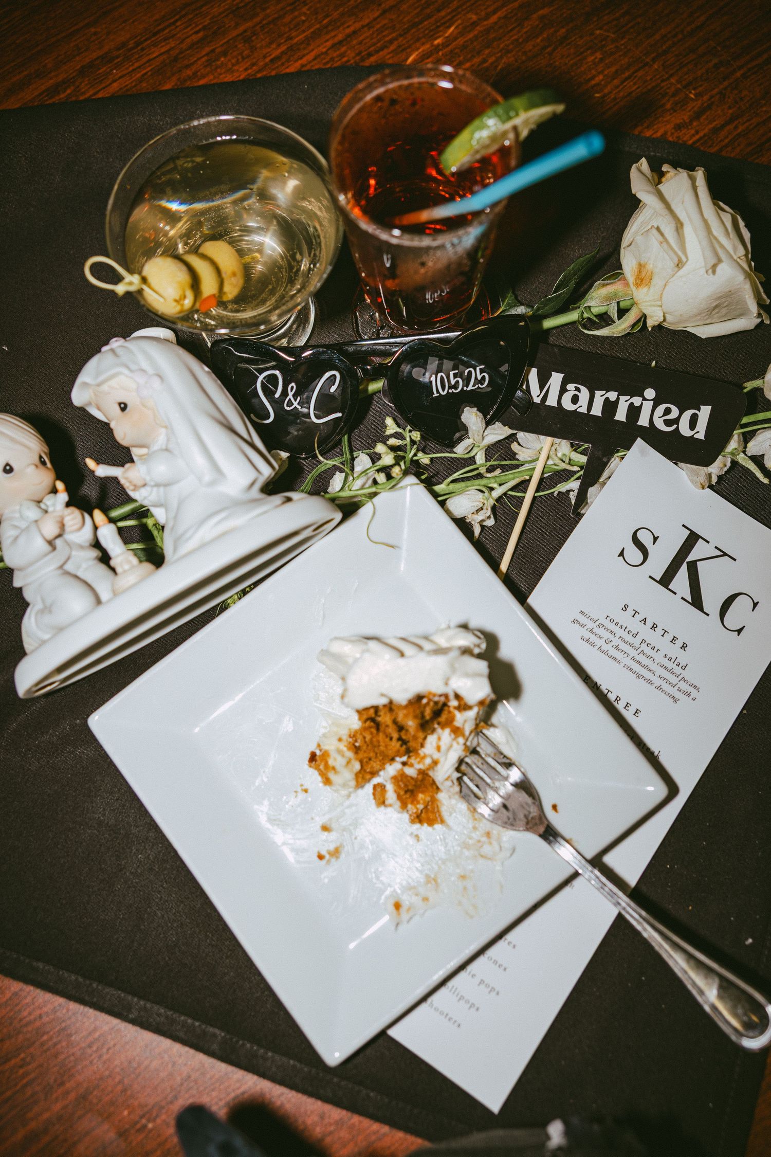 Wedding table flatlay with cake slice, cocktails, Just Married sign, and couple figurines on dark tablecloth.
