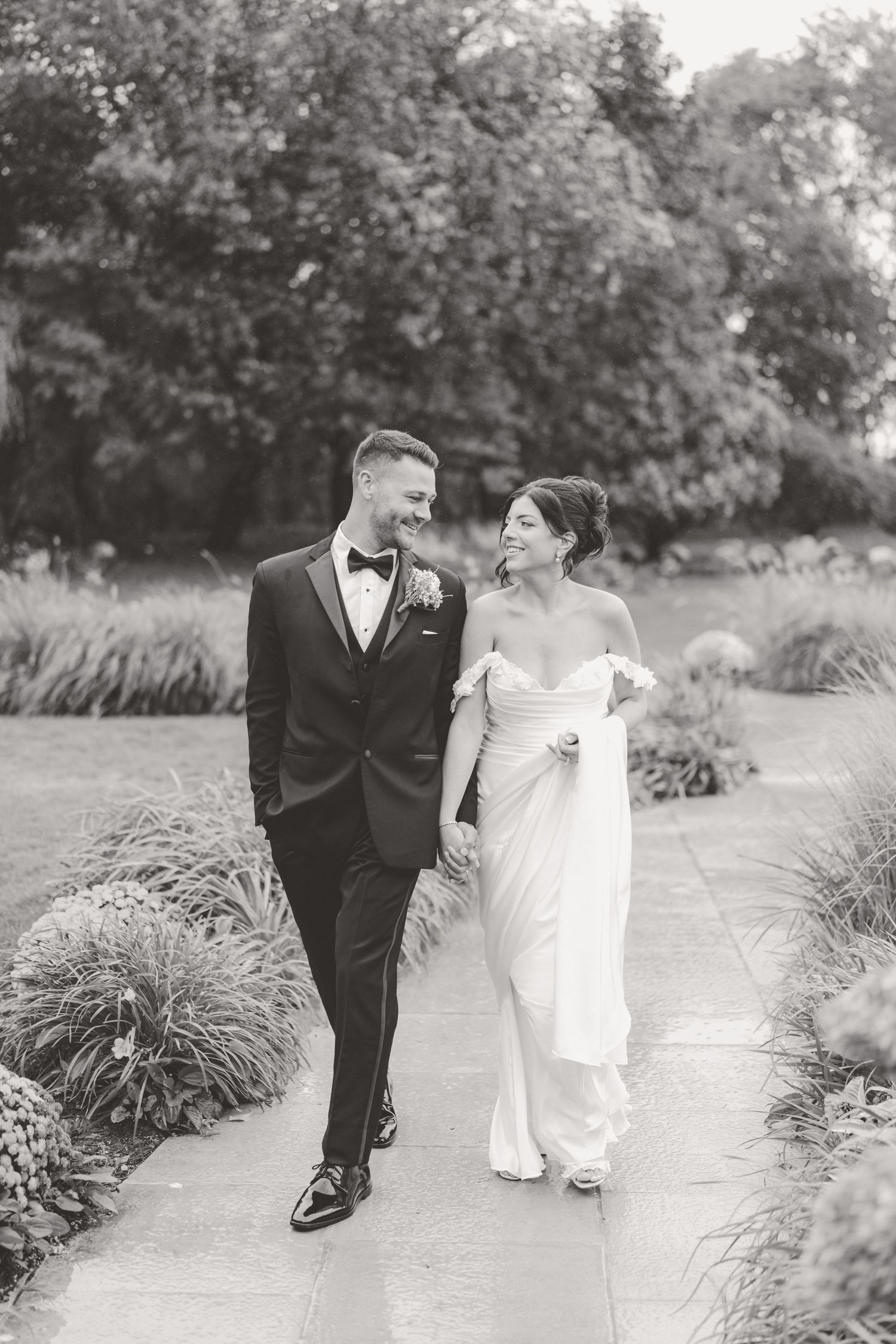 Bride and groom walk hand in hand along a garden path, smiling at each other on their wedding day.