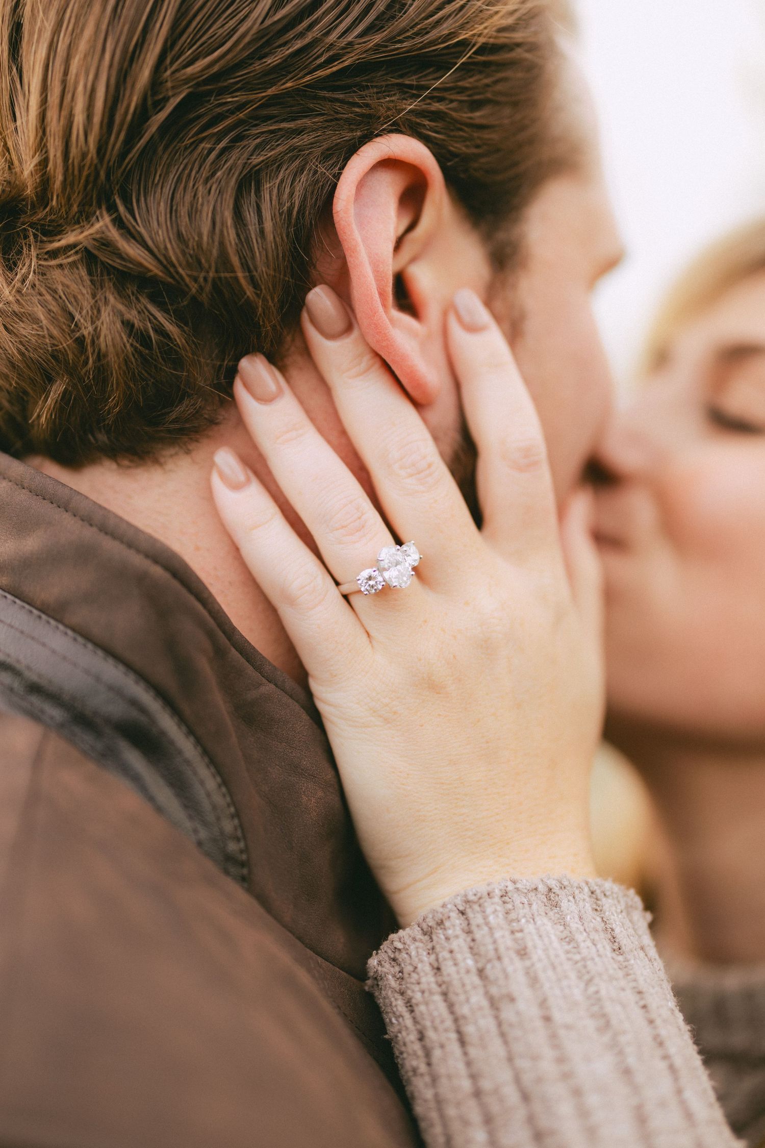 Woman wearing a three-stone diamond engagement ring tenderly kissing a man's cheek outdoors.
