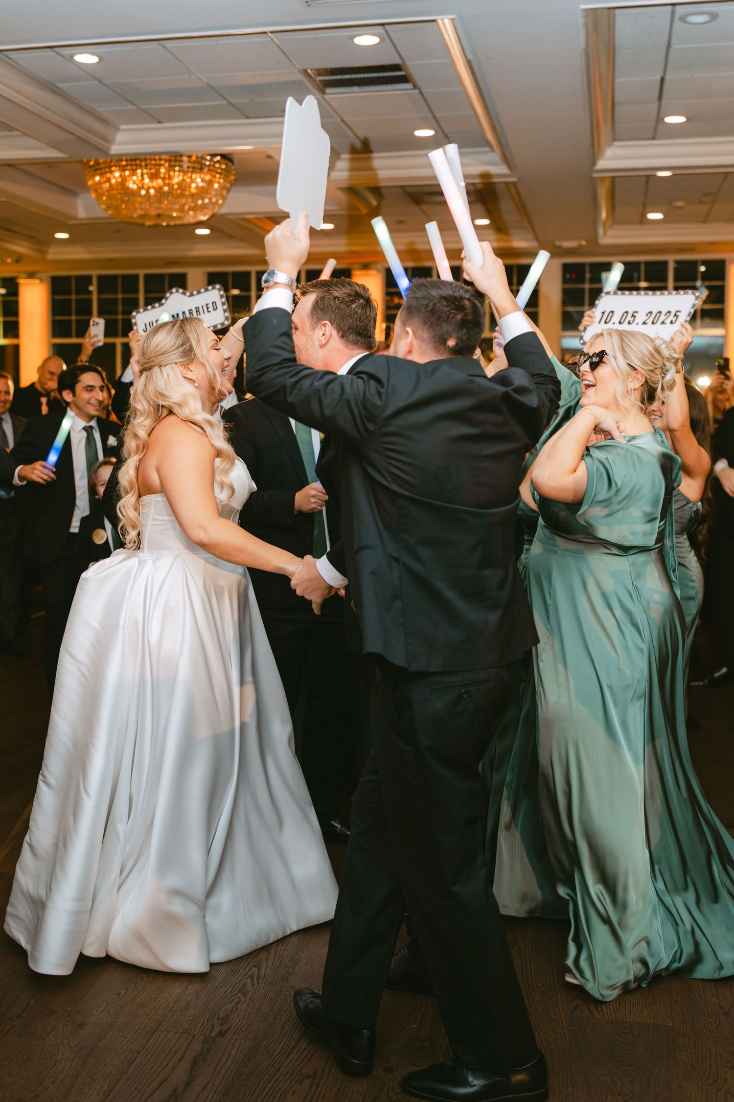 Bride and groom celebrate on the dance floor with guests holding glow sticks at lively wedding reception.
