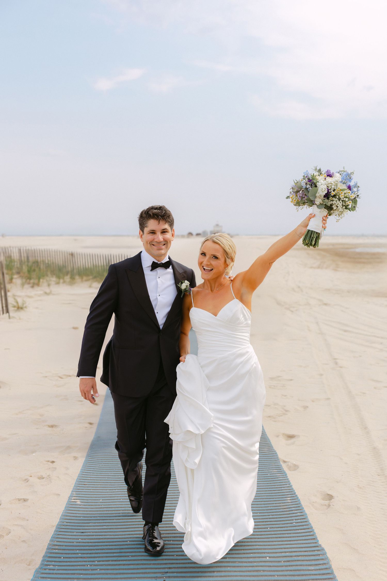 Elated bride raises bouquet of purple flowers while walking with groom on sunny beach boardwalk.
