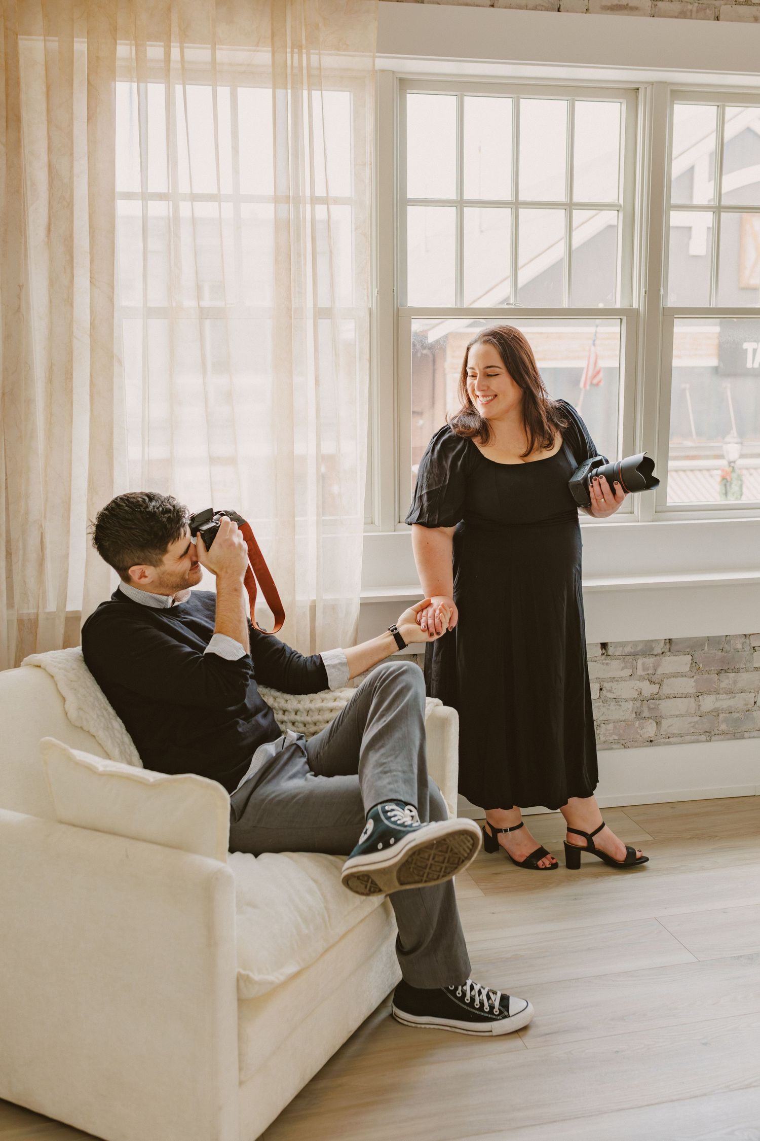Male photographer captures female colleague smiling in bright airy studio with large windows.