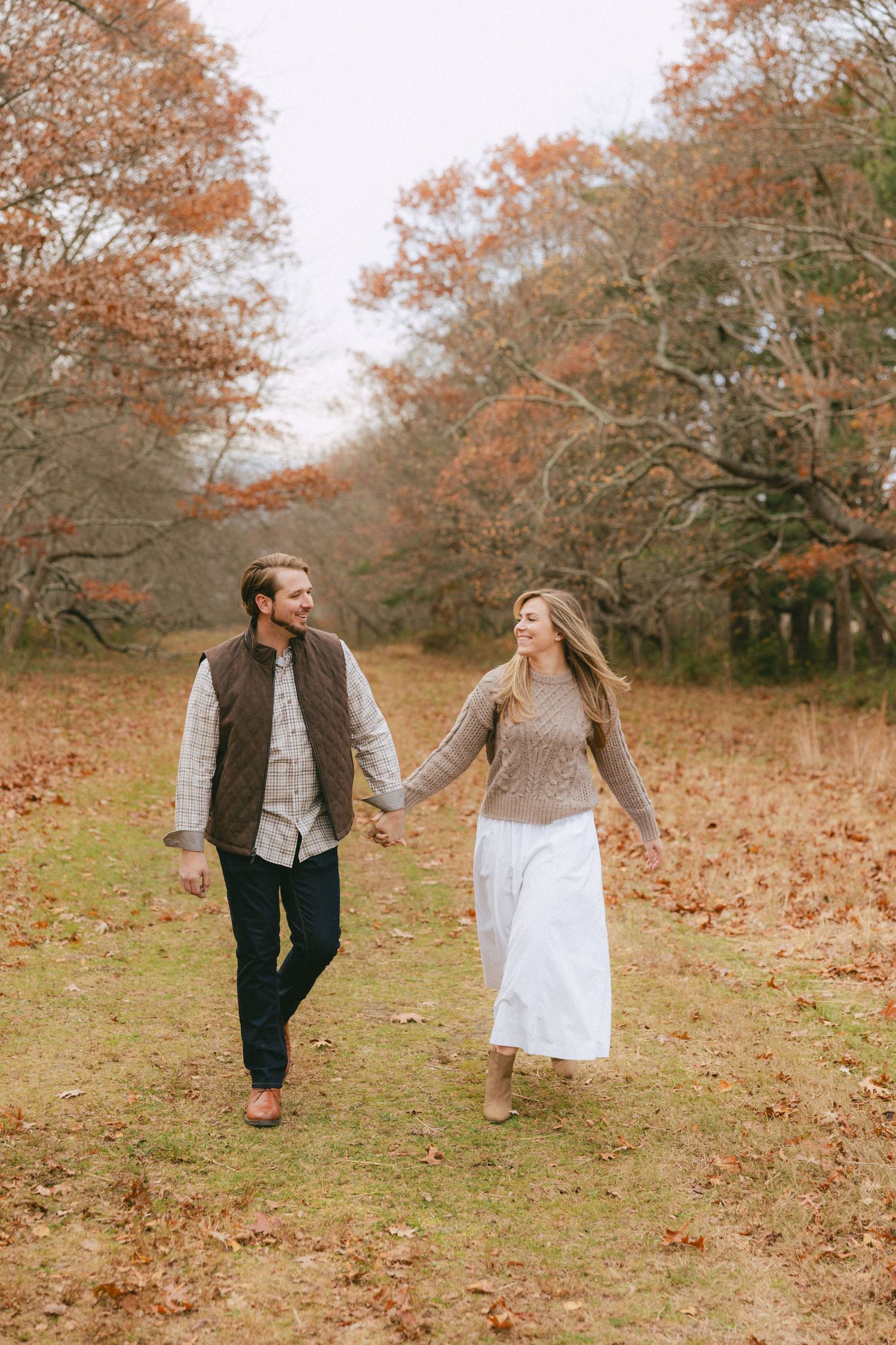 Happy couple holding hands and walking through an open autumn meadow with colorful fall trees.