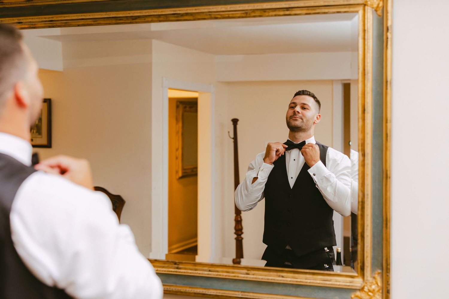 Groom adjusting bow tie while looking at his reflection in a large gold-framed mirror.
