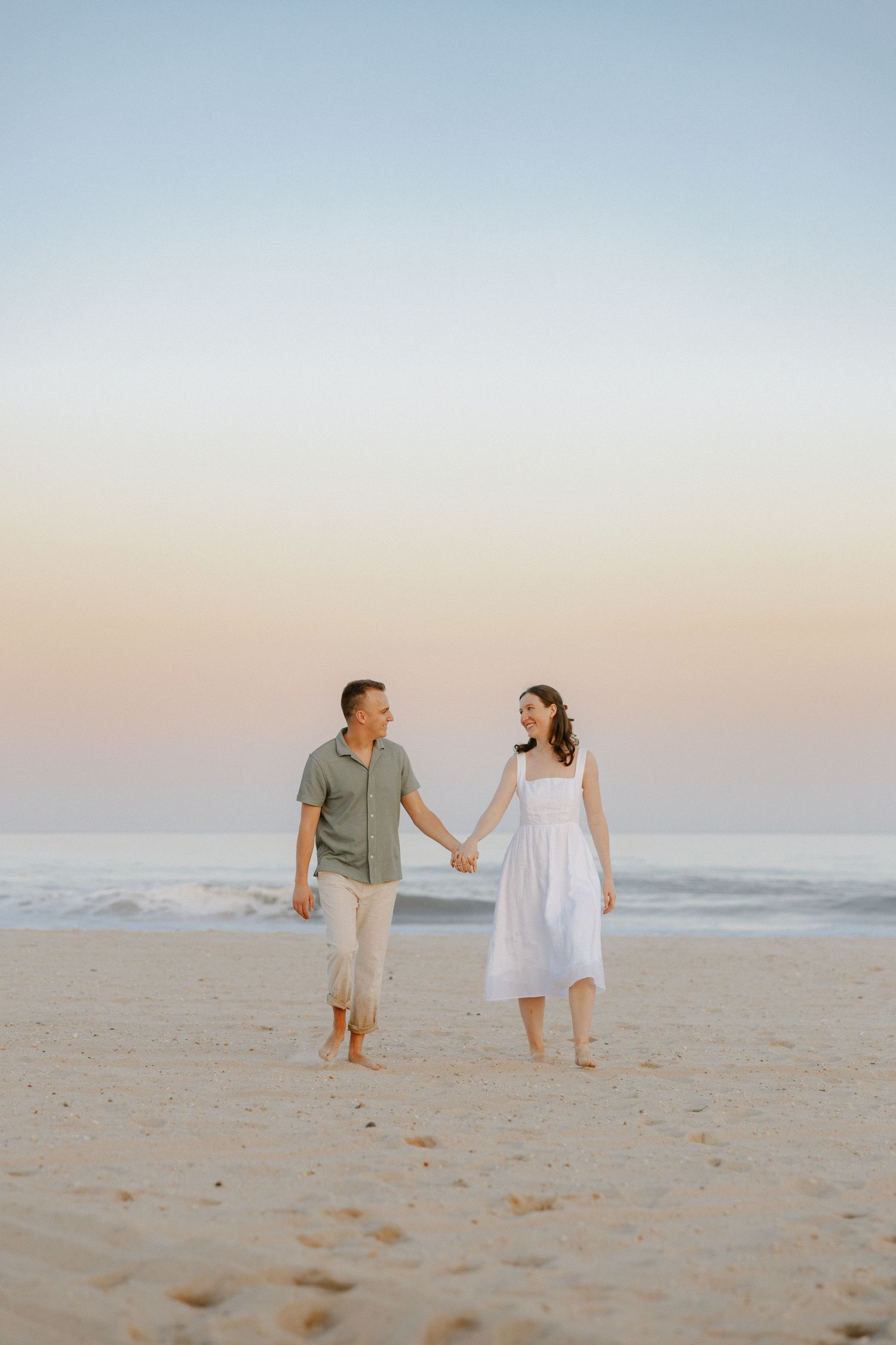 Couple holding hands and walking barefoot on beach at twilight with pastel sunset sky backdrop.