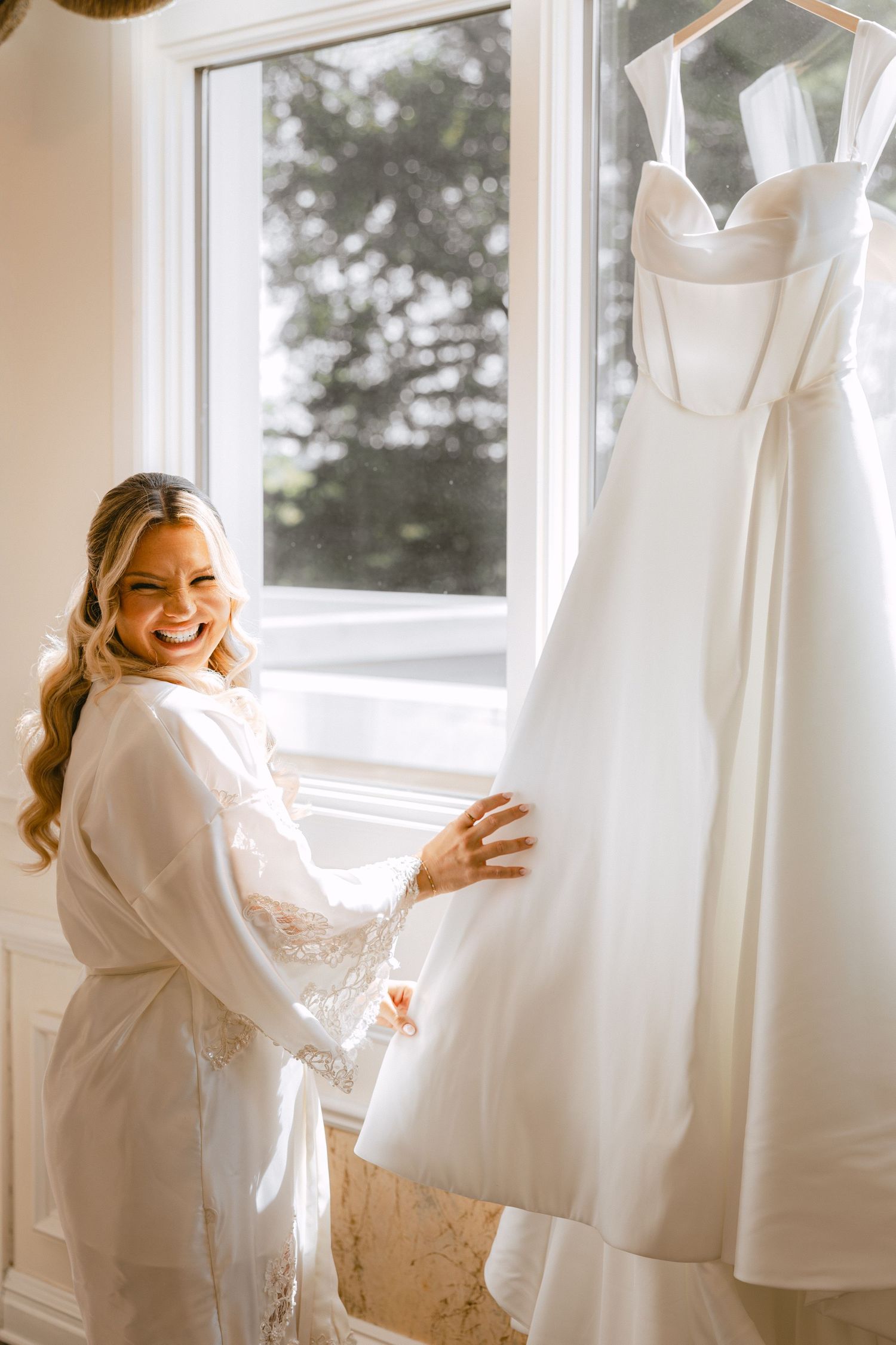 Radiant bride in lace robe smiling joyfully beside her elegant white wedding gown hanging in window.