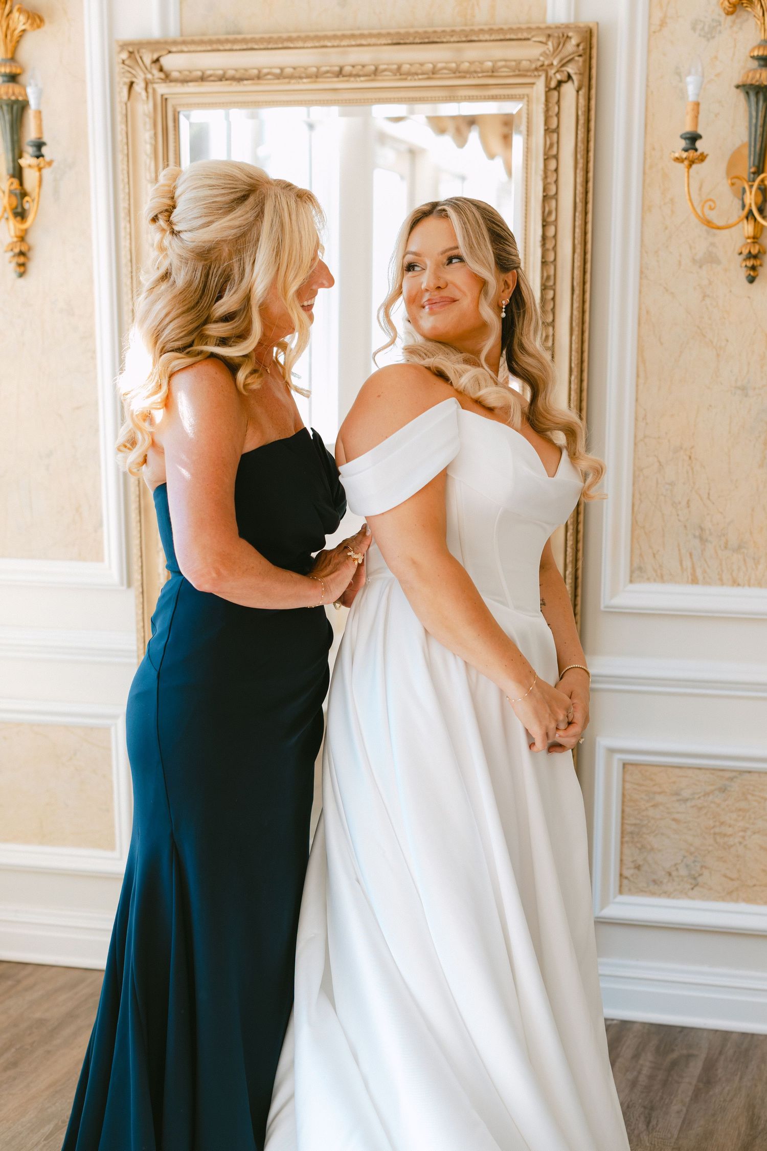 Bride in off-shoulder white gown smiling with bridesmaid in navy dress in front of ornate gold mirror.