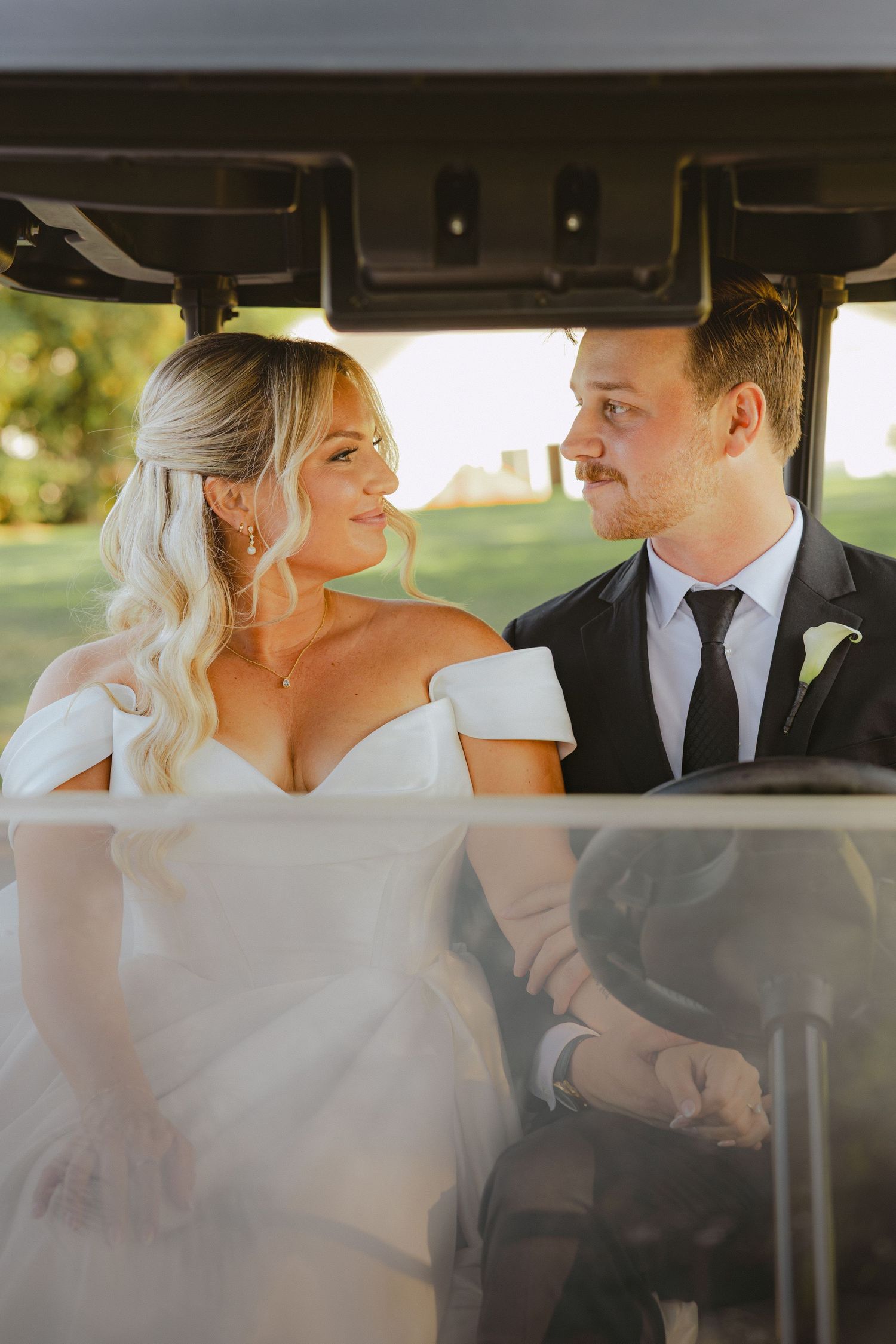 Bride and groom gaze lovingly at each other while seated together in a golf cart at sunset.