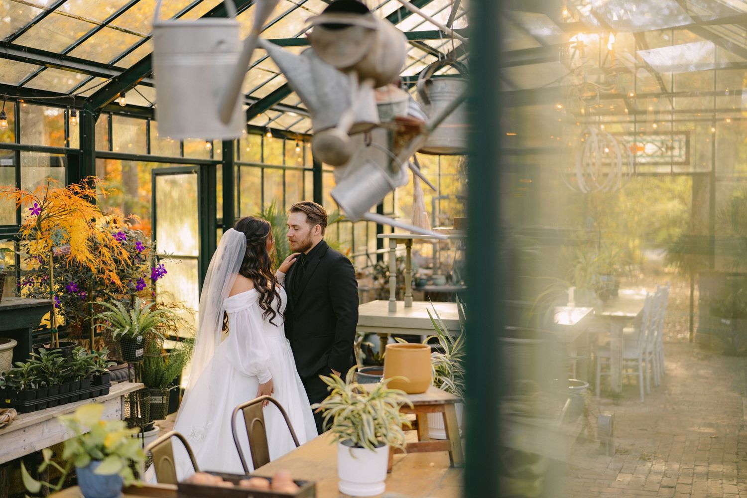 Bride and groom share a romantic moment inside a sunlit greenhouse surrounded by lush plants and flowers.