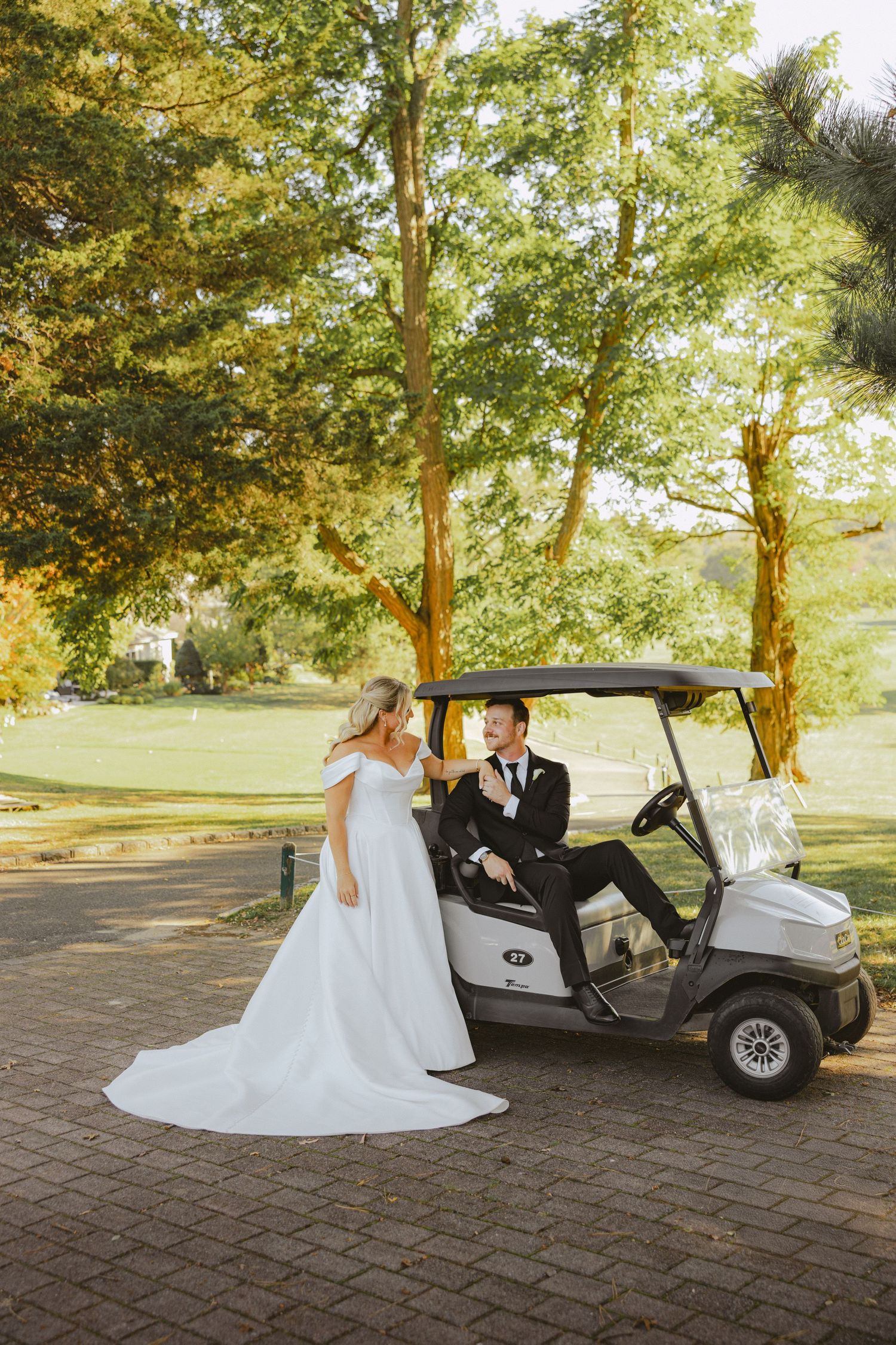 Groom sits on a golf cart reaching toward his bride in a sunlit tree-lined outdoor venue setting.