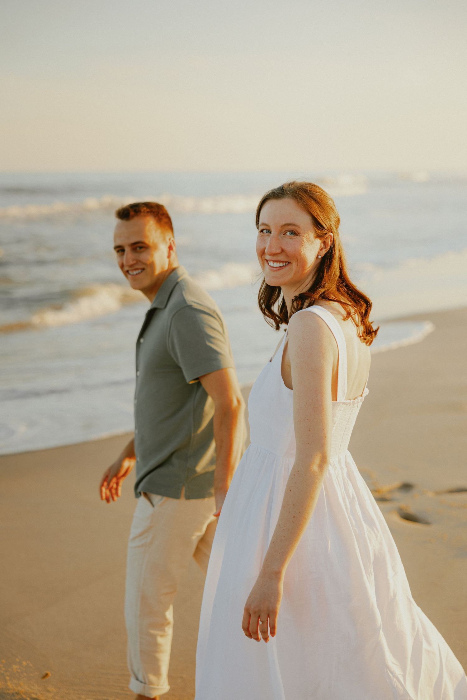 Smiling redhead bride in white dress walks with groom on beach at warm sunset.